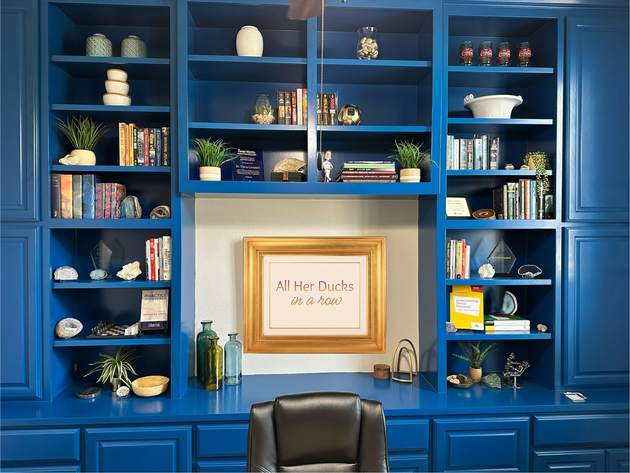 A blue built-in bookshelf filled with books, decorative objects, plants, rocks, and glass bottles, with a framed sign that reads, "All Her Ducks in a Row," in a room with a gray wall and a black chair in the foreground.