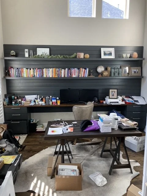 Home office with a dark wall unit holding books, framed photos, and decorative items; cluttered desk with papers, books, and office supplies; beige office chair; window letting in natural light; various boxes and items on the floor.