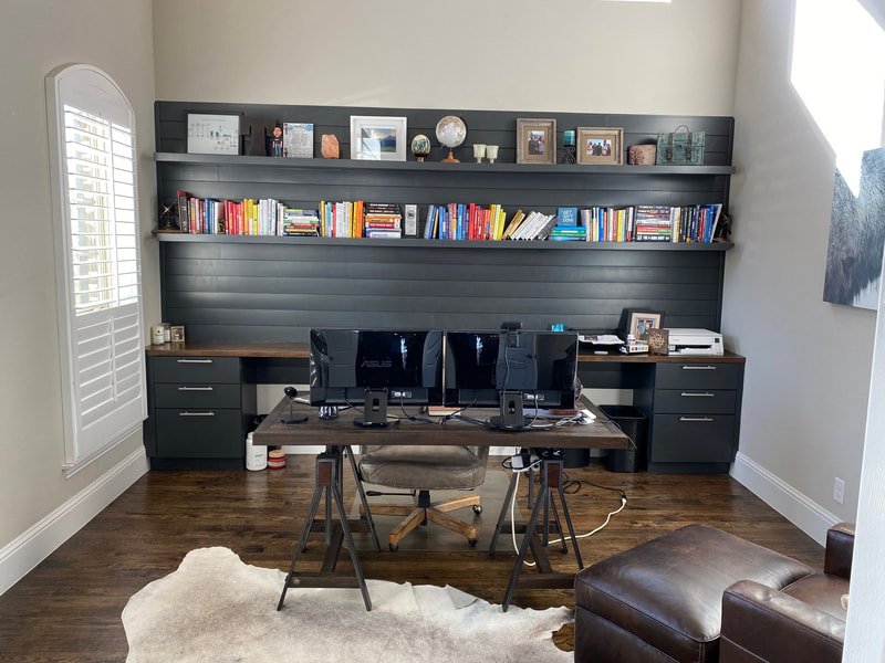 Home office with dark wood desk, two monitors, and chairs, set against a wall with two shelves filled with books and decorative items, a window with white shutters, and a leather chair in the foreground.