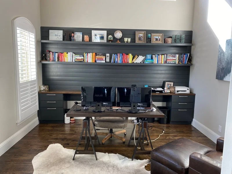 Home office with a desk, three computer monitors, and a laptop; dark wood shelving with books and decorative items; gray walls and hardwood floor.