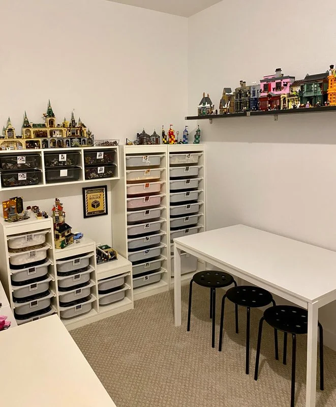 A room with white walls and carpeted floor, containing multiple white storage units filled with LEGO bricks, some displayed as LEGO castles on top. There is a white table with three black stools and a shelf with colorful LEGO models on the wall.