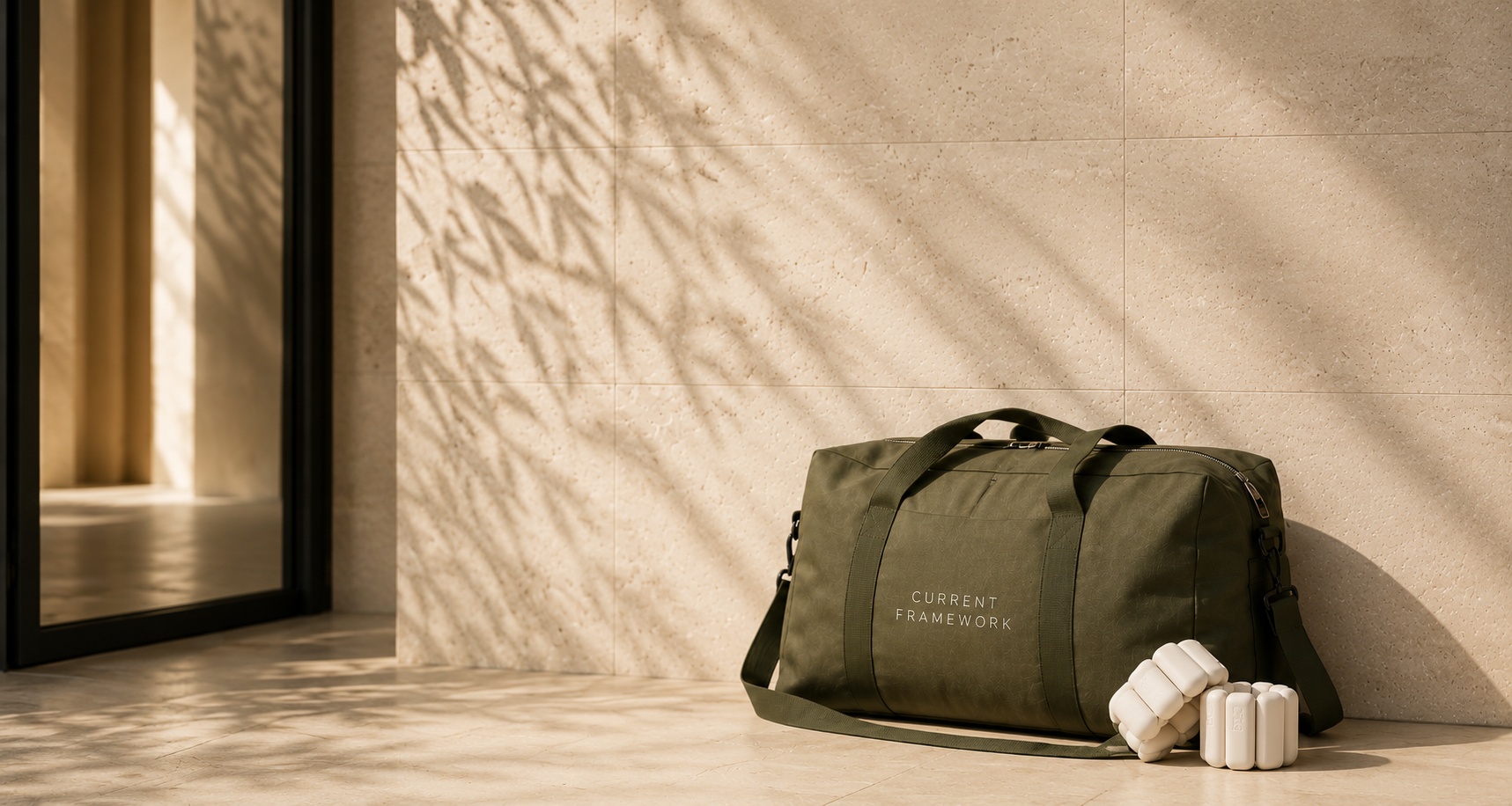 A green duffel bag labeled 'CURRENT FRAMEWORK' on the side, resting against a beige tiled wall, with weighted pilates bangles in front of it. Sunlight creates shadows of plants on the wall.