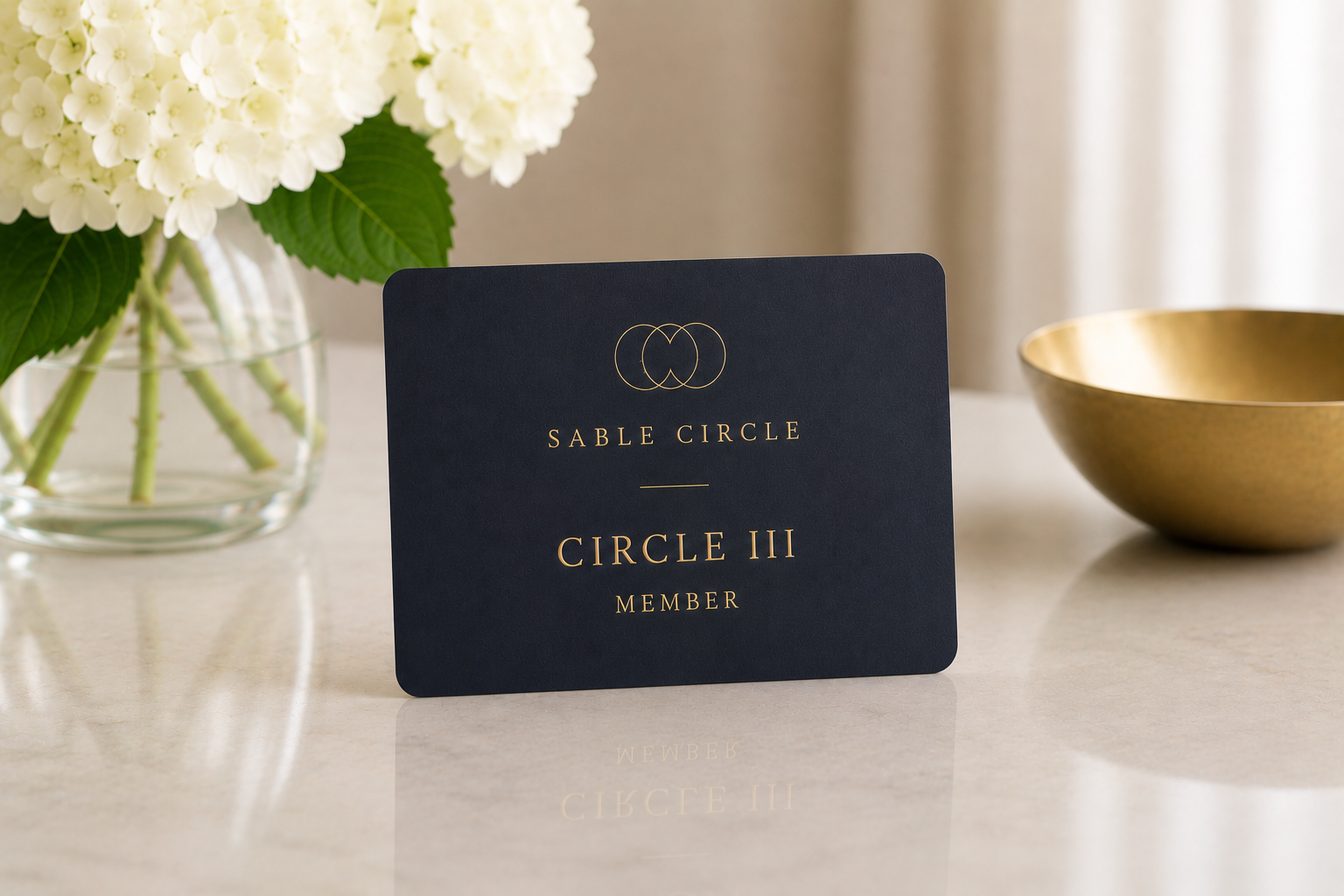 A navy blue card with gold text and logo that reads "Sable Circle," "Circle III," and "Member" on a light-colored surface. There is a glass vase with white hydrangea flowers and a green leaf to the left, and a gold-colored bowl to the right.