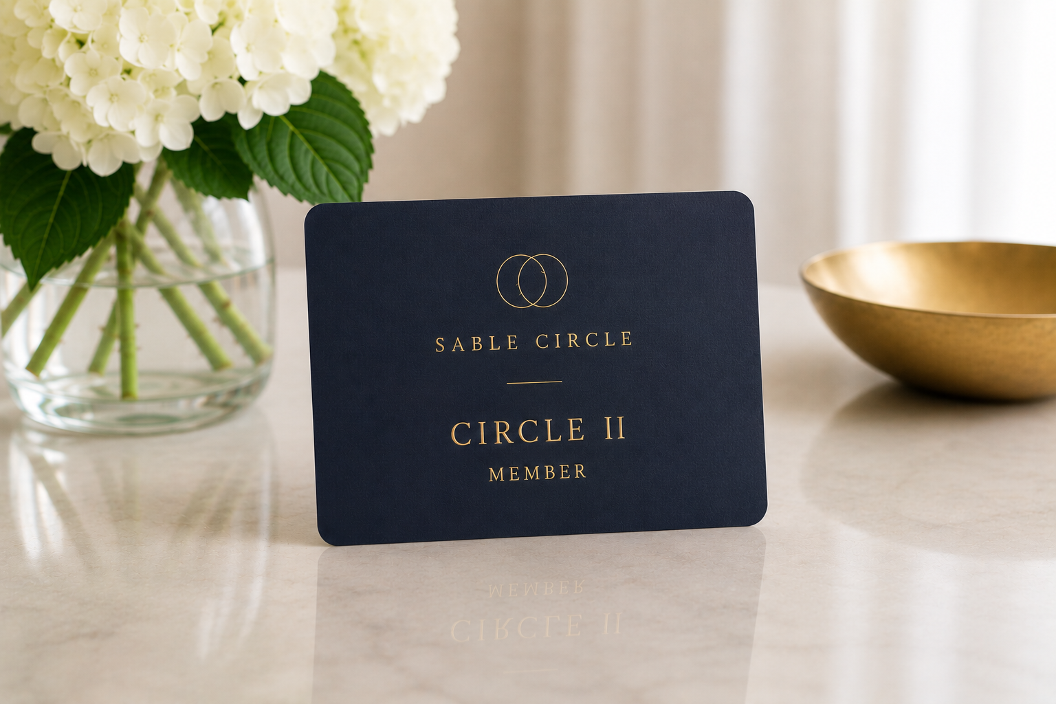 A navy blue card with gold text and circles reading 'Sable Circle,' 'Circle II,' and 'Member,' placed on a light-colored surface. To the left, a glass vase with white hydrangea flowers, and to the right, a gold bowl.