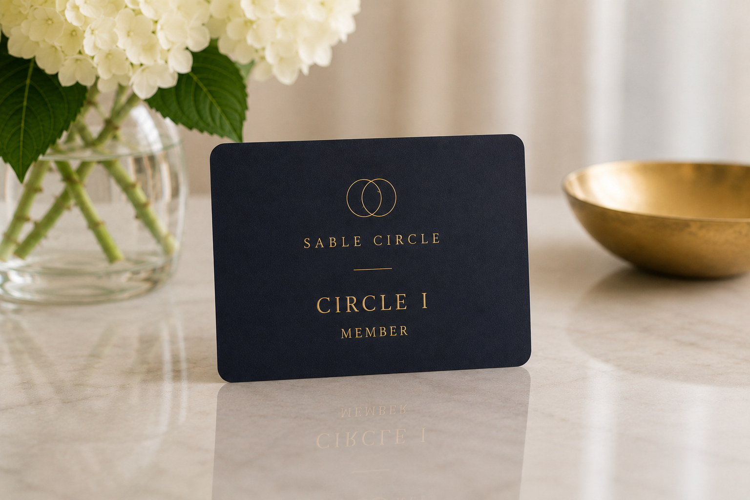 A black membership card with gold foil lettering that reads "Sable Circle Circle 1 Member" in a serif font, placed on a polished white marble surface. To the left, there is a clear glass vase with white hydrangea flowers and green leaves. To the right, there is a gold bowl.