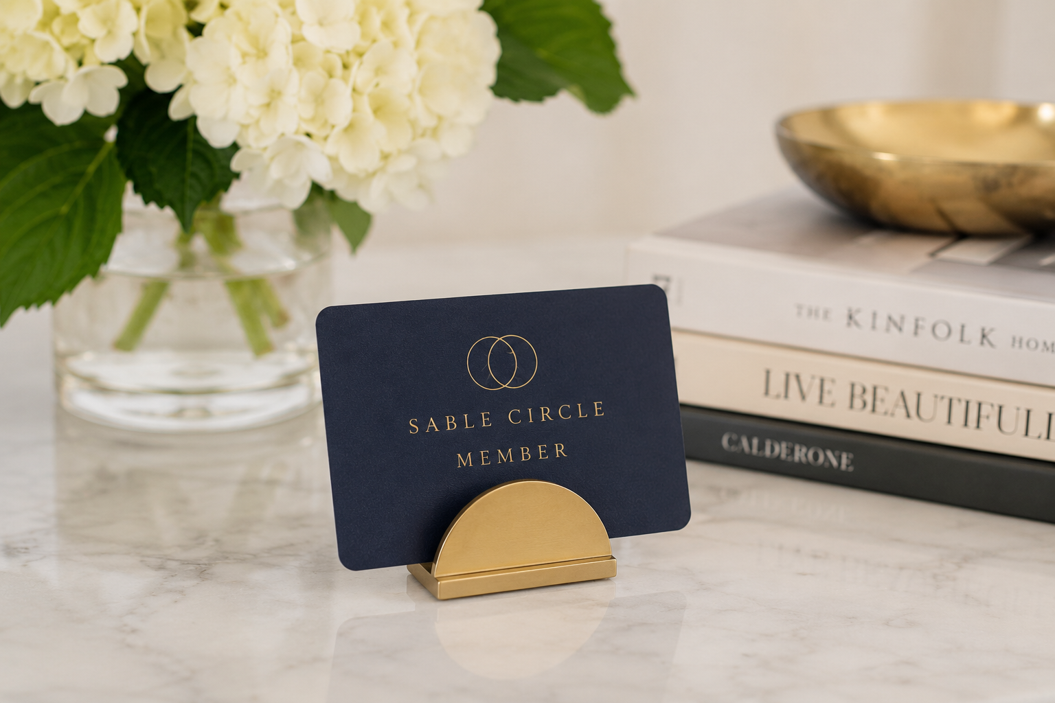 A navy blue Sable Circle Member card with gold lettering and a gold circle logo, displayed on a gold stand on a white marble surface. In the background, there is a glass vase with white hydrangeas, and stacked hardcover books, with a gold bowl on top.