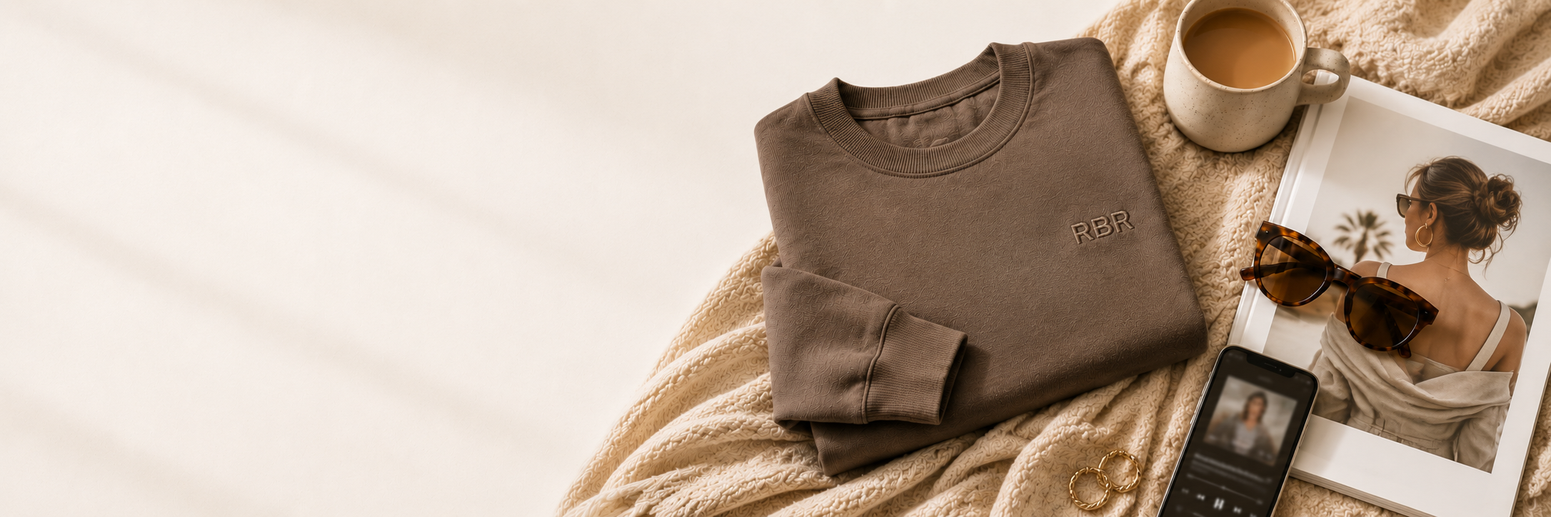 Flat lay of beige blanket, folded brown sweatshirt with RBR logo, beige mug of coffee, sunglasses, a smartphone, a magazine with a photo of a woman with sunglasses and a towel on her shoulders, and a pair of gold hoop earrings.