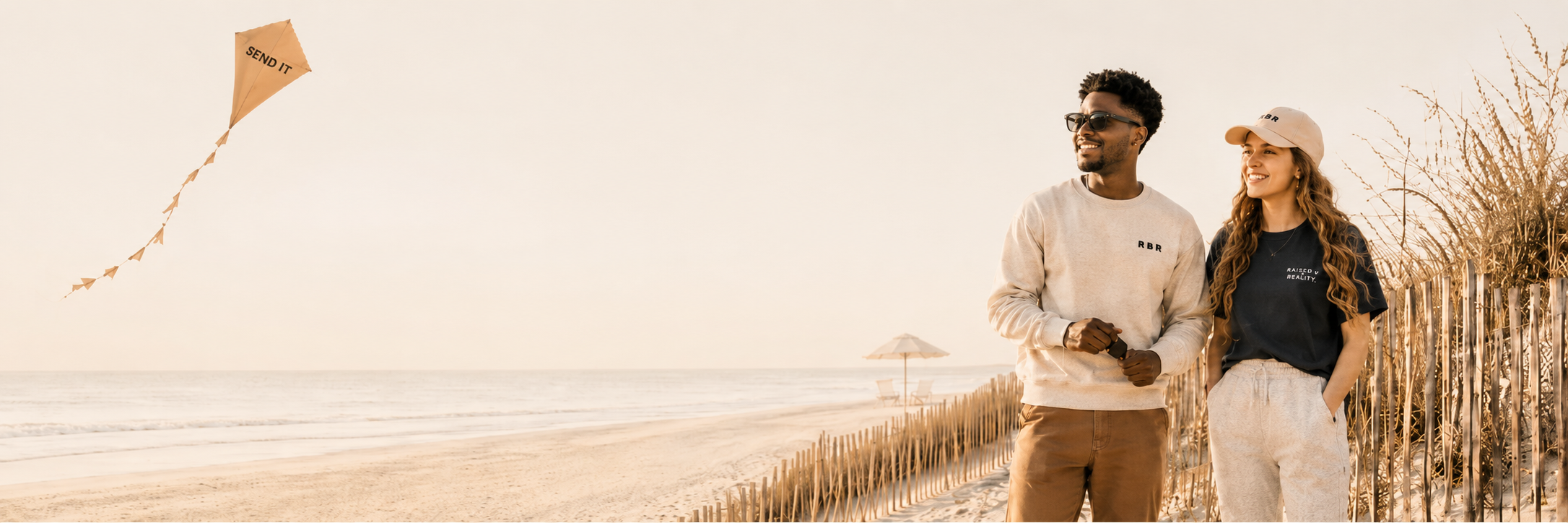 A young man and woman walking on a beach, smiling, with a kite flying in the sky, sand dunes, and beach umbrellas in the background.