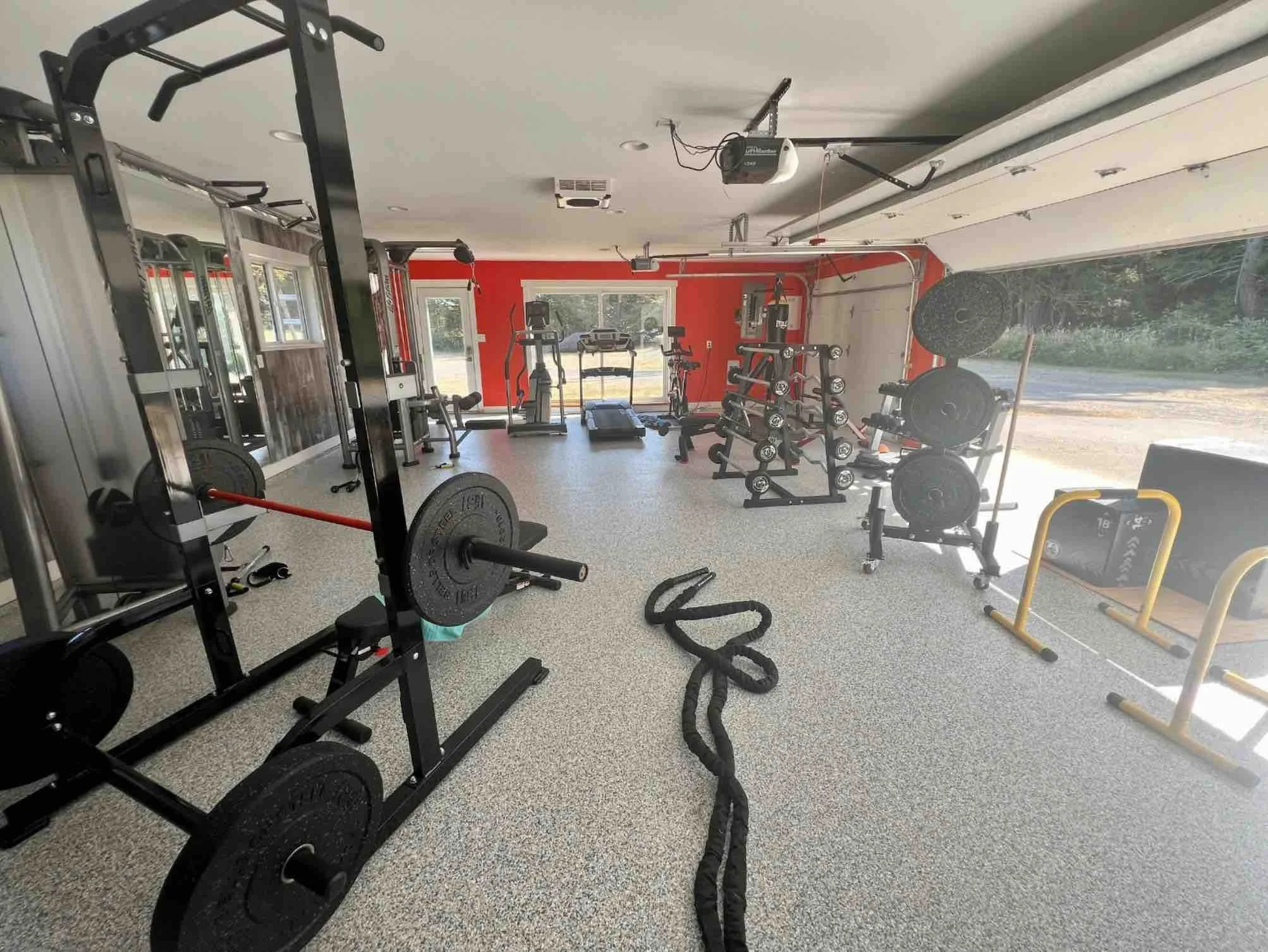 Home gym with various workout equipment including weights, a treadmill, a rowing machine, and a pull-up bar, with large windows allowing natural light.