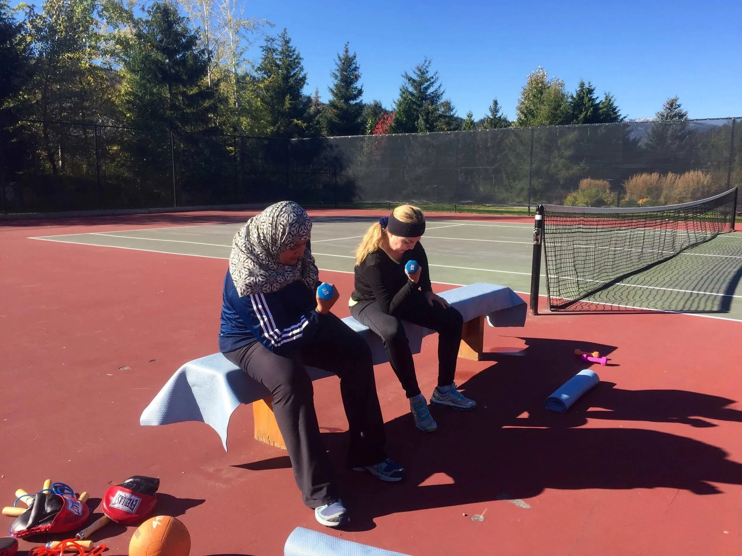 two women on the tennis court lifting weights, one wearing a hijab. it is sunny