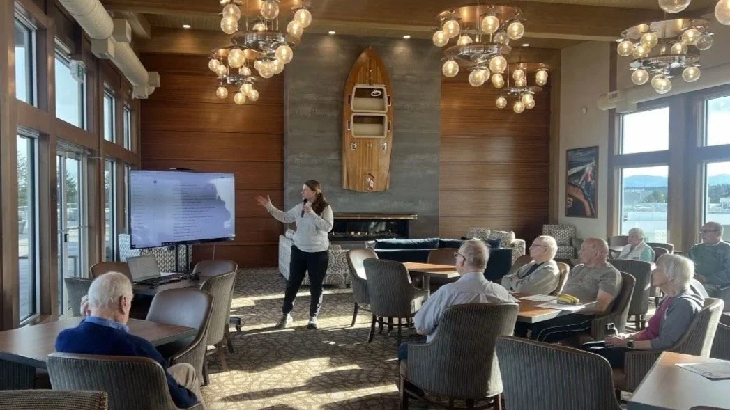 A woman giving a presentation to an audience in a well-lit room with large windows, wooden accent walls, a mounted TV, and decorative light fixtures hanging from the ceiling.