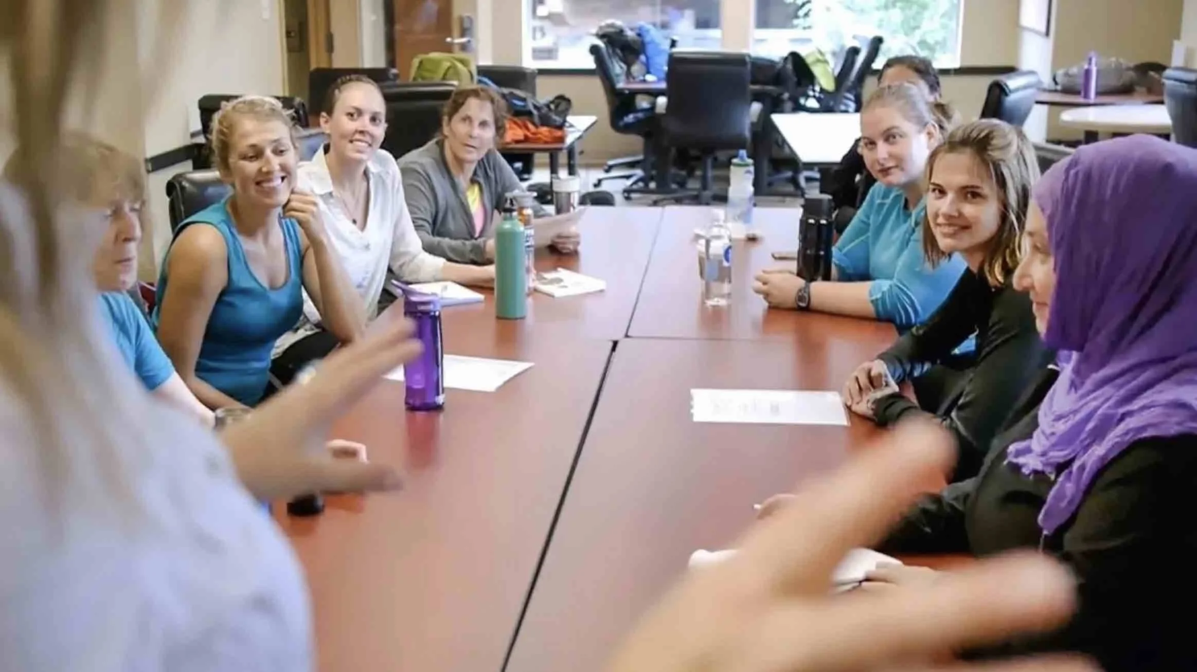 Group of women sitting around a conference table in a meeting room, some smiling and engaging in conversation.