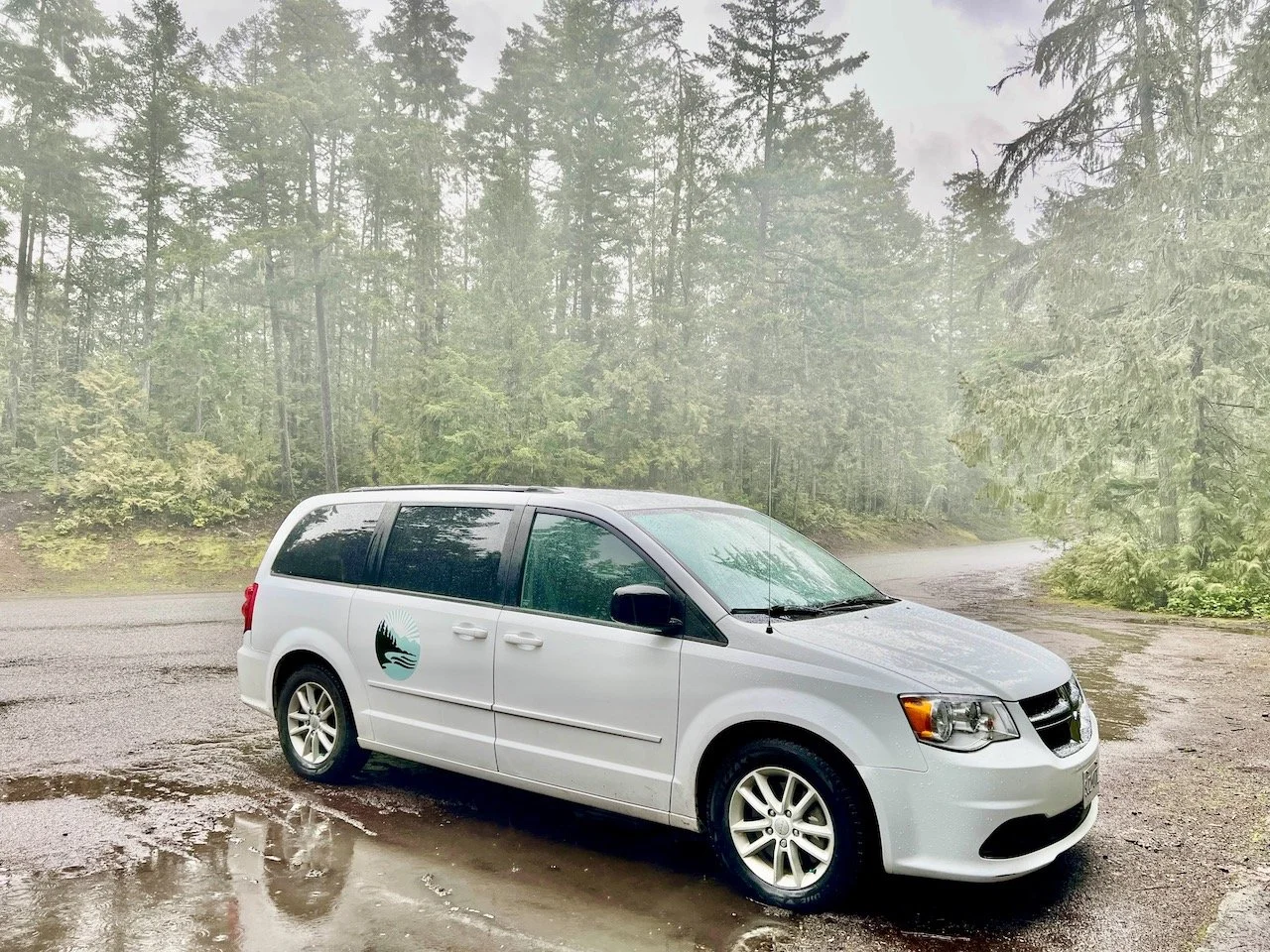 White minivan with a logo on the side parked on a wet dirt road in a forested area with tall trees.
