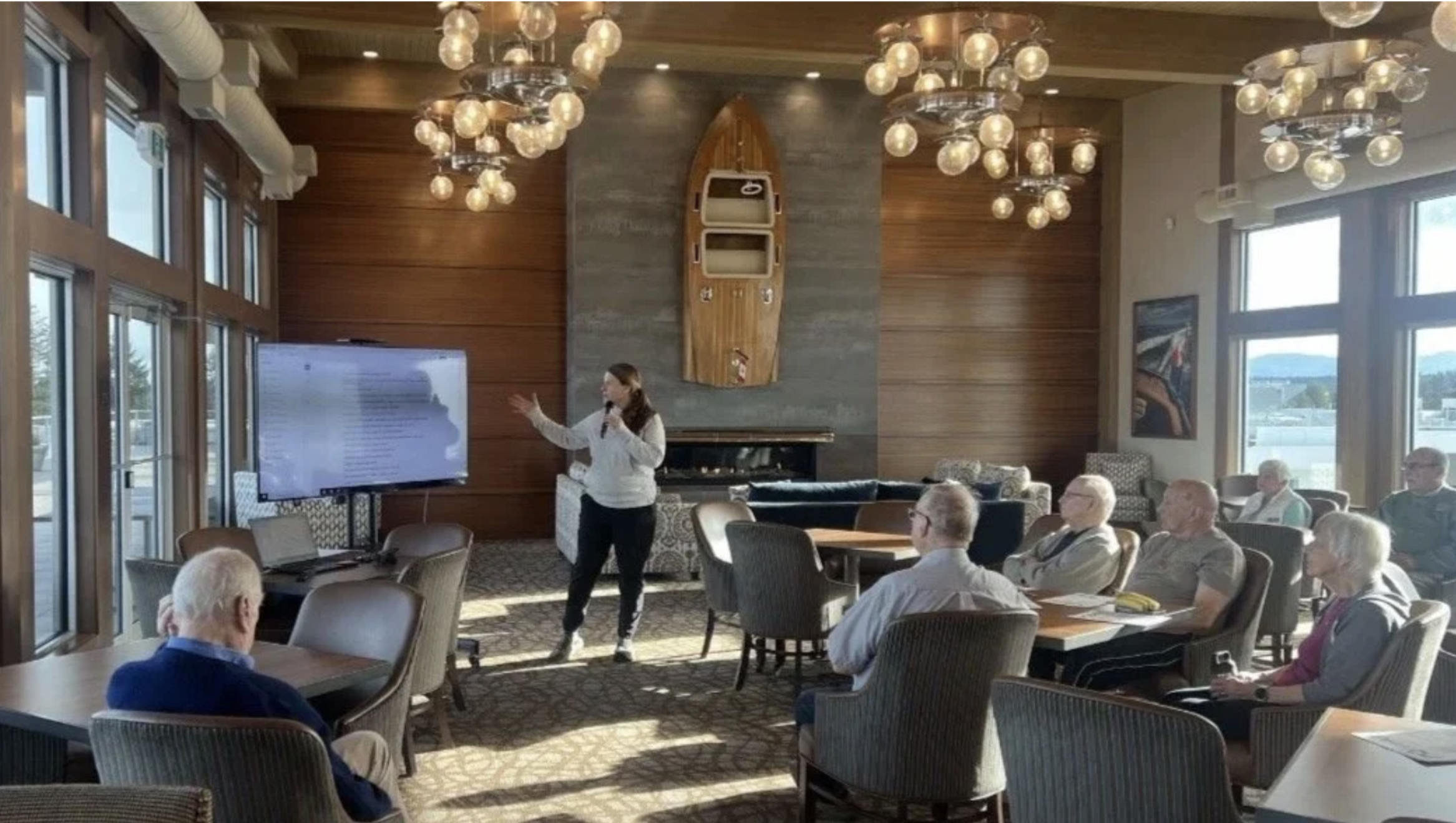 A woman giving a presentation to a group of elderly people in a well-lit room with wooden and stone walls, chandeliers, large windows, and framed artwork.