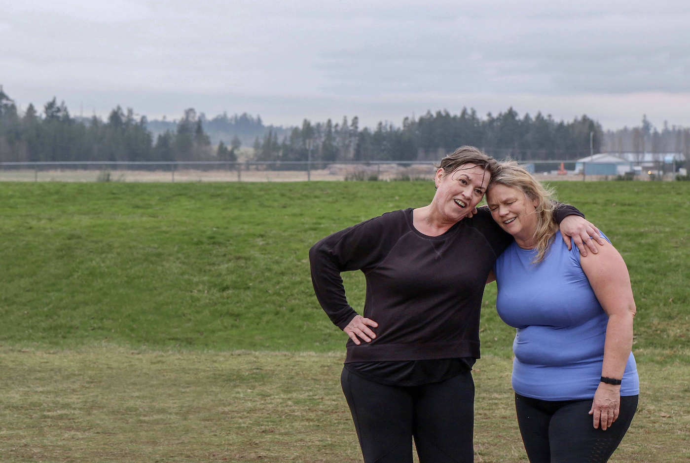Two women standing outdoors on a grassy field, smiling and leaning on each other, with cloudy skies and trees in the background.