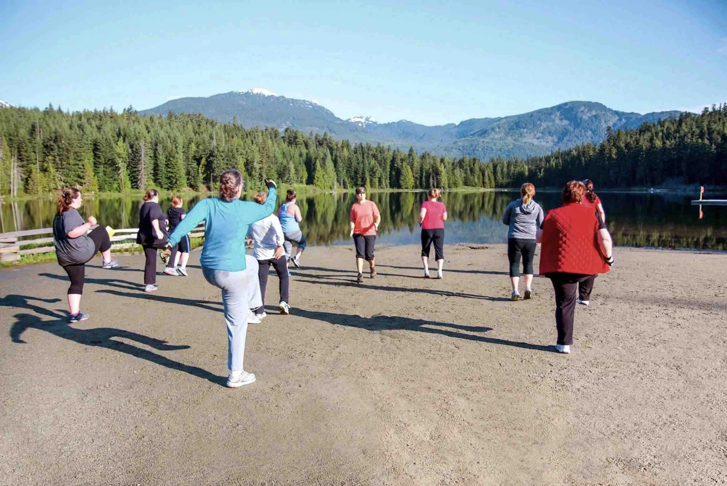Group of people practicing fitness in a scenic outdoor area with a lake, forest, and mountains in the background.