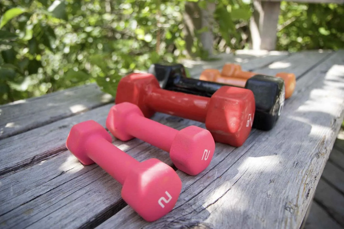 Two pink dumbbells and two red, black, and orange hand weights on a weathered wooden outdoor surface with green foliage in the background.