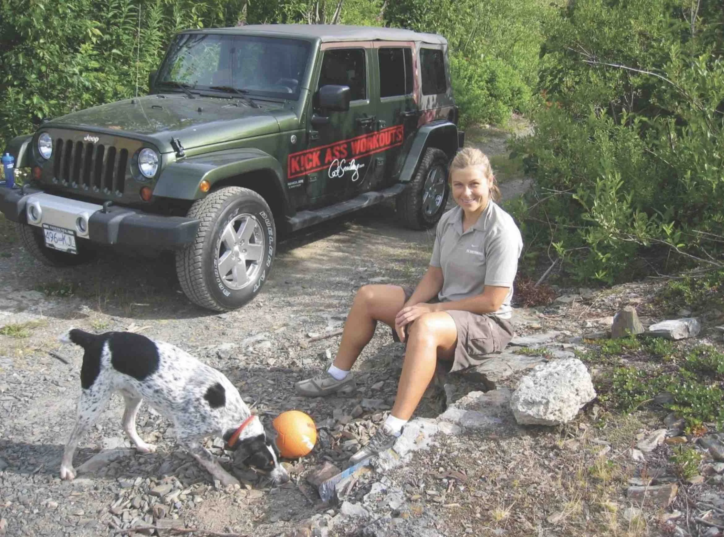 A woman with long blonde hair sitting on a rocky ground next to a black and white dog with spots, playing with an orange ball. Behind her is a green Jeep with a red and black sticker that says 'KICK ASS WORKOUTS' and a signature below it. The Jeep is parked on a dirt path surrounded by dense green foliage.
