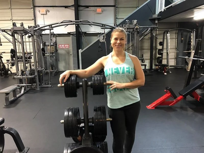 A woman in athletic wear smiling at the camera in a gym, standing next to a weight rack with dumbbells, surrounded by gym equipment.