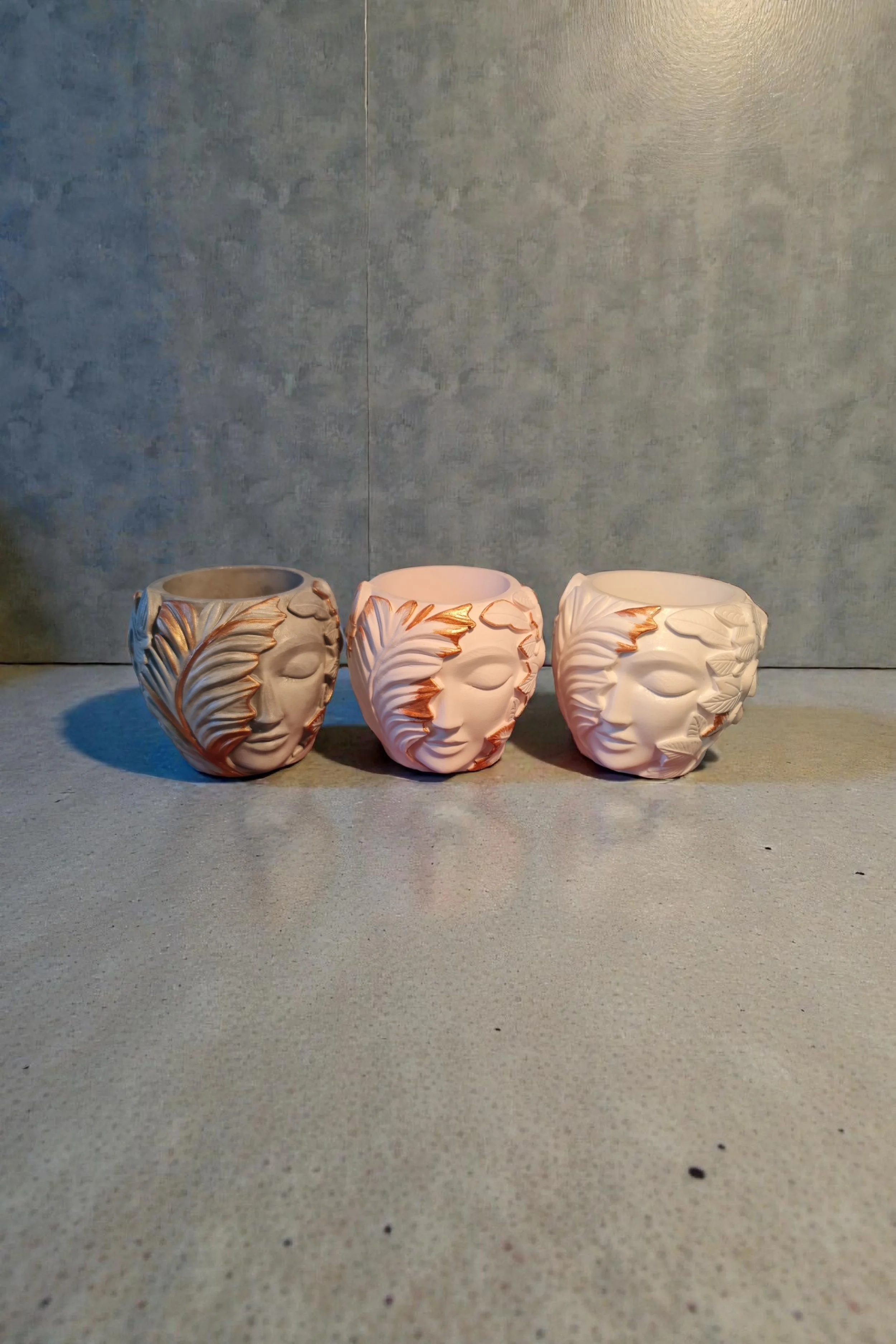 Three ceramic cups designed with faces and gold accents, arranged on a flat surface against a gray wall background.