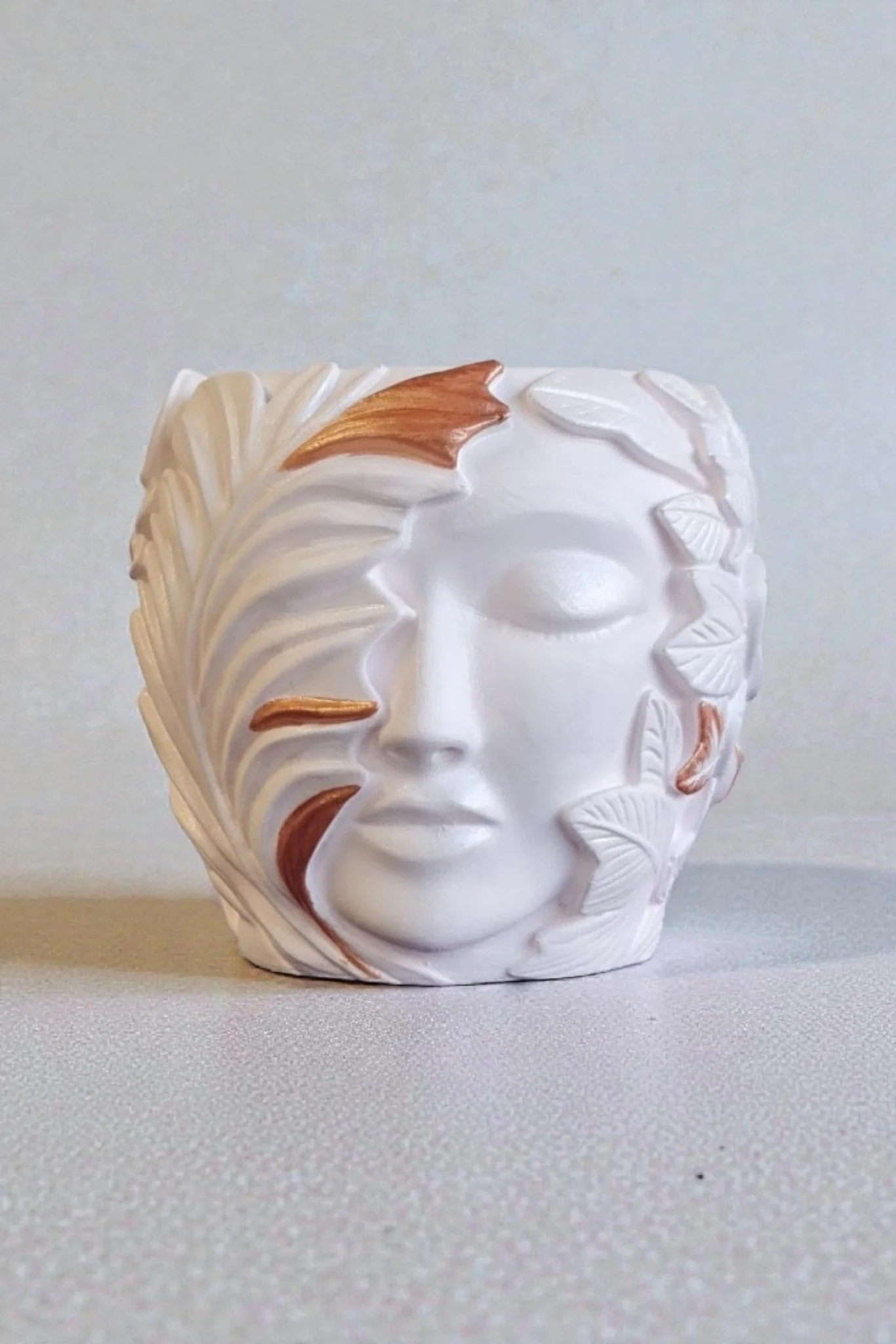 A white Gypsum Cement Vessel, with hand-painted detail leaf and palm frond designs, accented with gold mica, sitting on a light-colored surface with a plain light background.