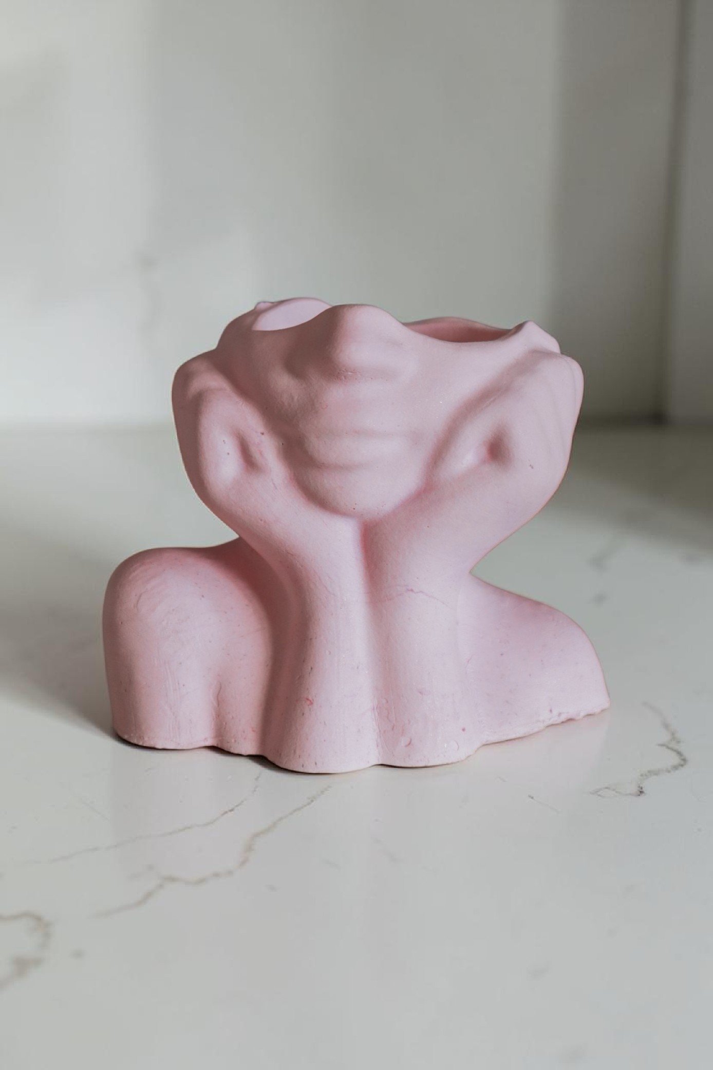 Pink abstract sculpture resembling a human face with closed eyes, set on a white surface with subtle marbling.