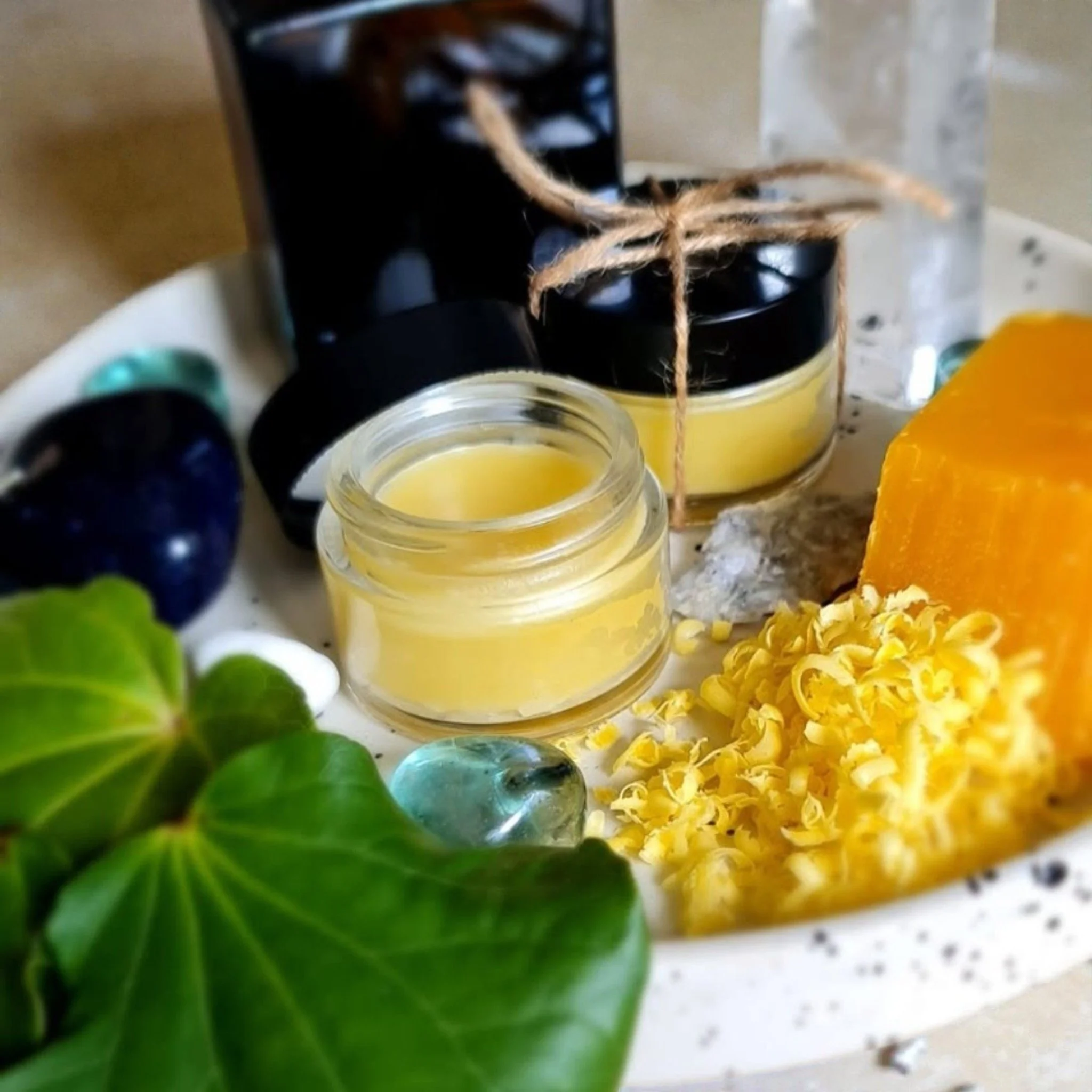 Close-up of small glass jars filled with yellow beeswax, tied with twine, surrounded by natural ingredients such as green leaves and blackberries, on a white speckled surface.
