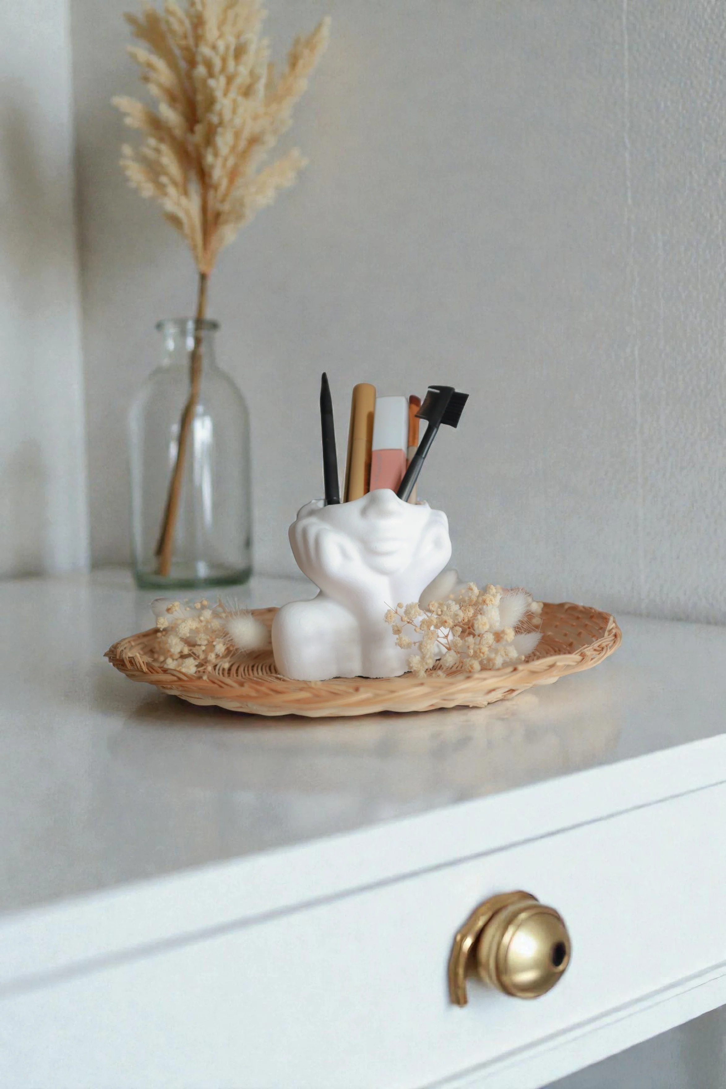 A white dresser with a gold knob, a wicker tray, a white ceramic container with pens inside, a small vase with dried pampas grass, and a dollop of white cream or mousse with flowers on top.