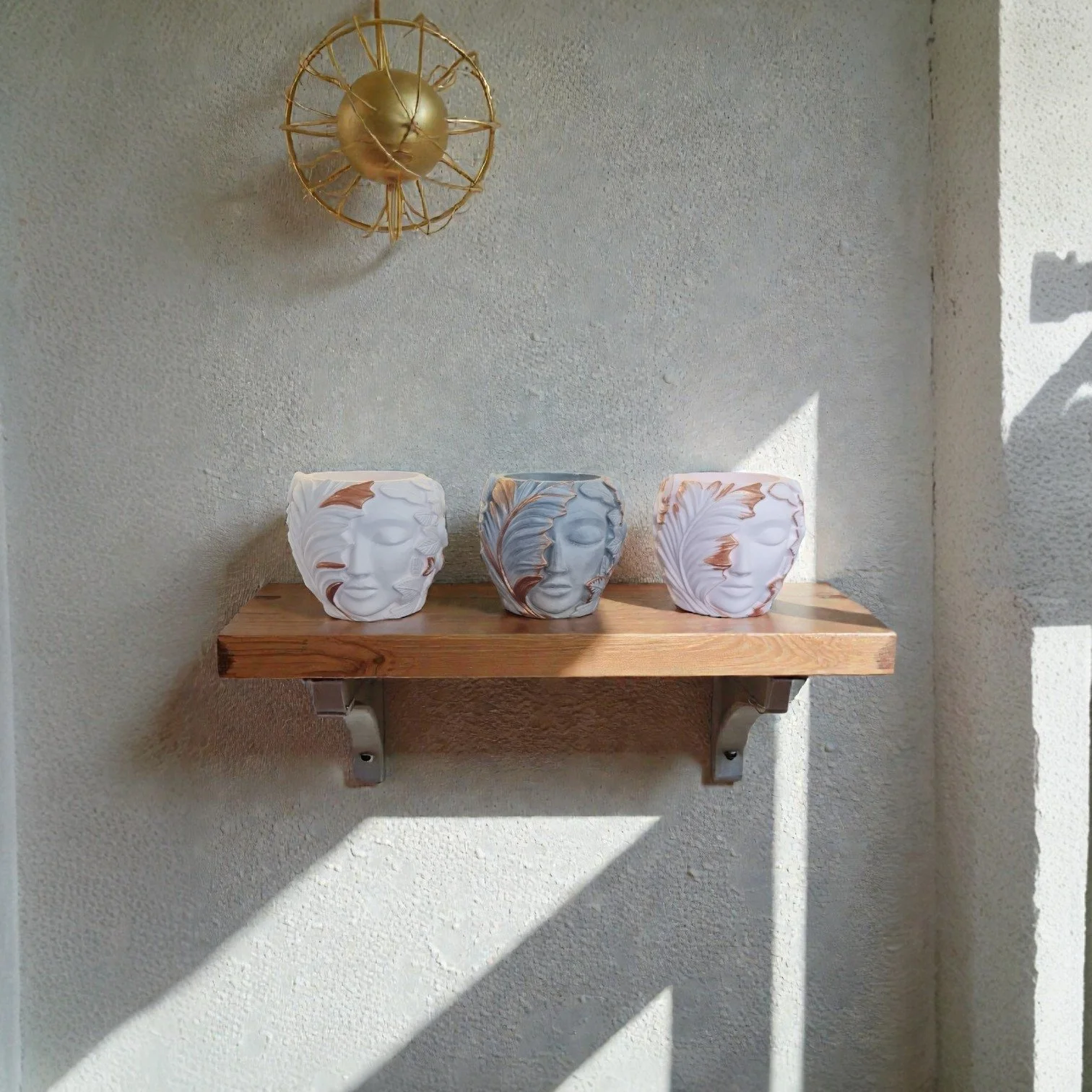 Three ceramic plant pots with carved female faces, one white, one gray, and one white with brown accents, on a wooden shelf mounted on a beige wall with sunlight and shadows.