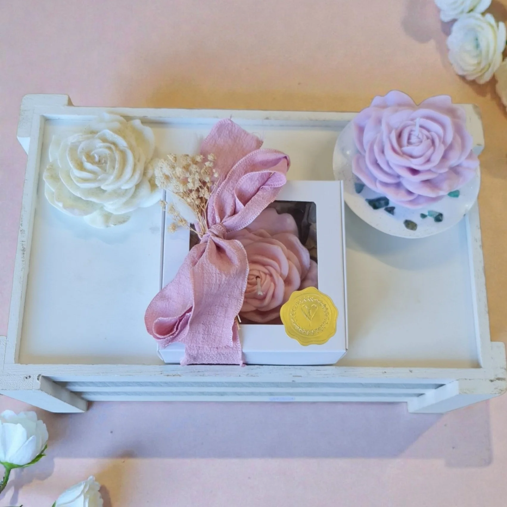 Decorative white and pink roses, a small bouquet of dried flowers, a pink fabric ribbon, and a yellow wax seal on a white wooden tray