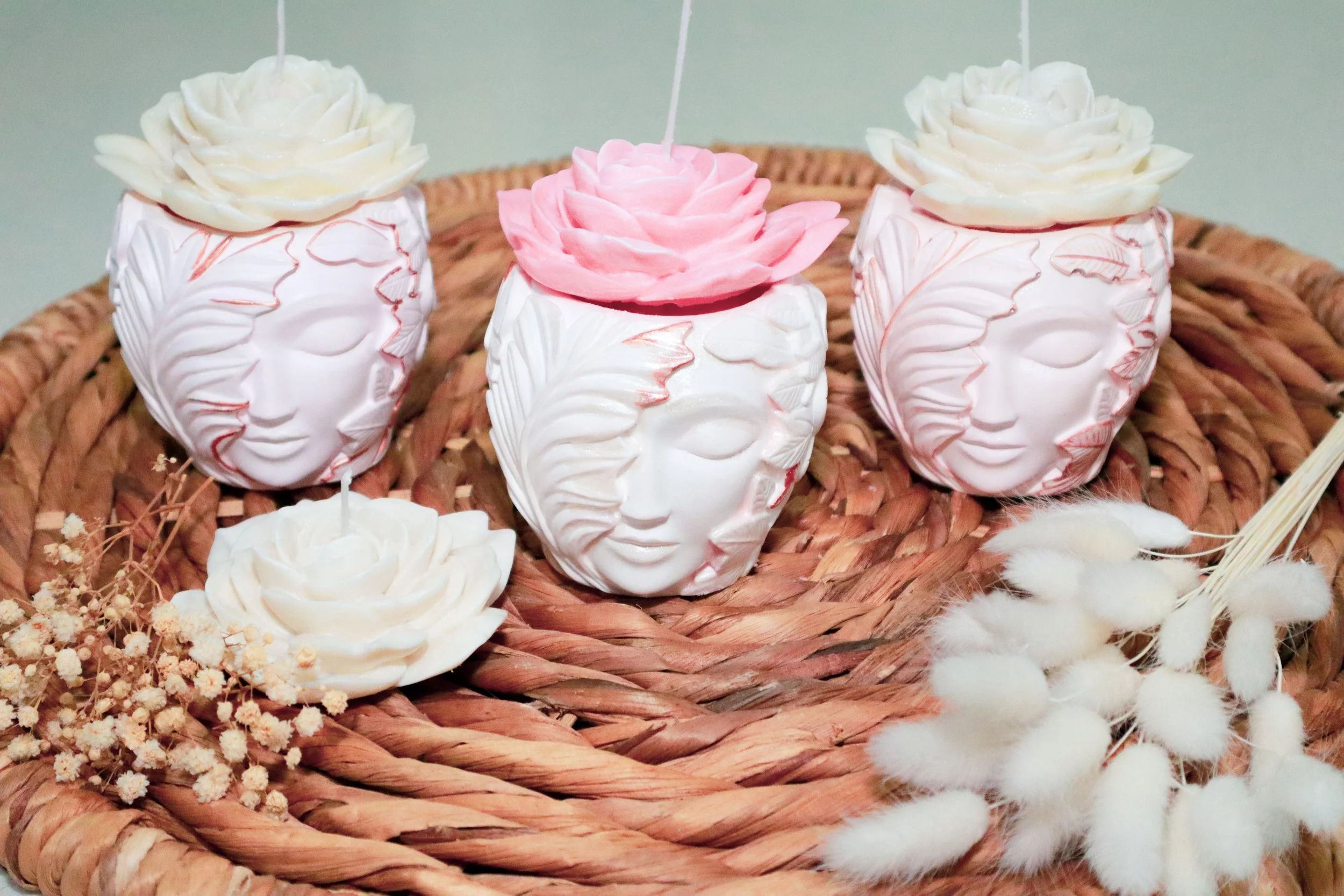 Decorative white and pink flower-shaped candles in carved white holders on a woven basket with white and pink accents and cotton sprigs.