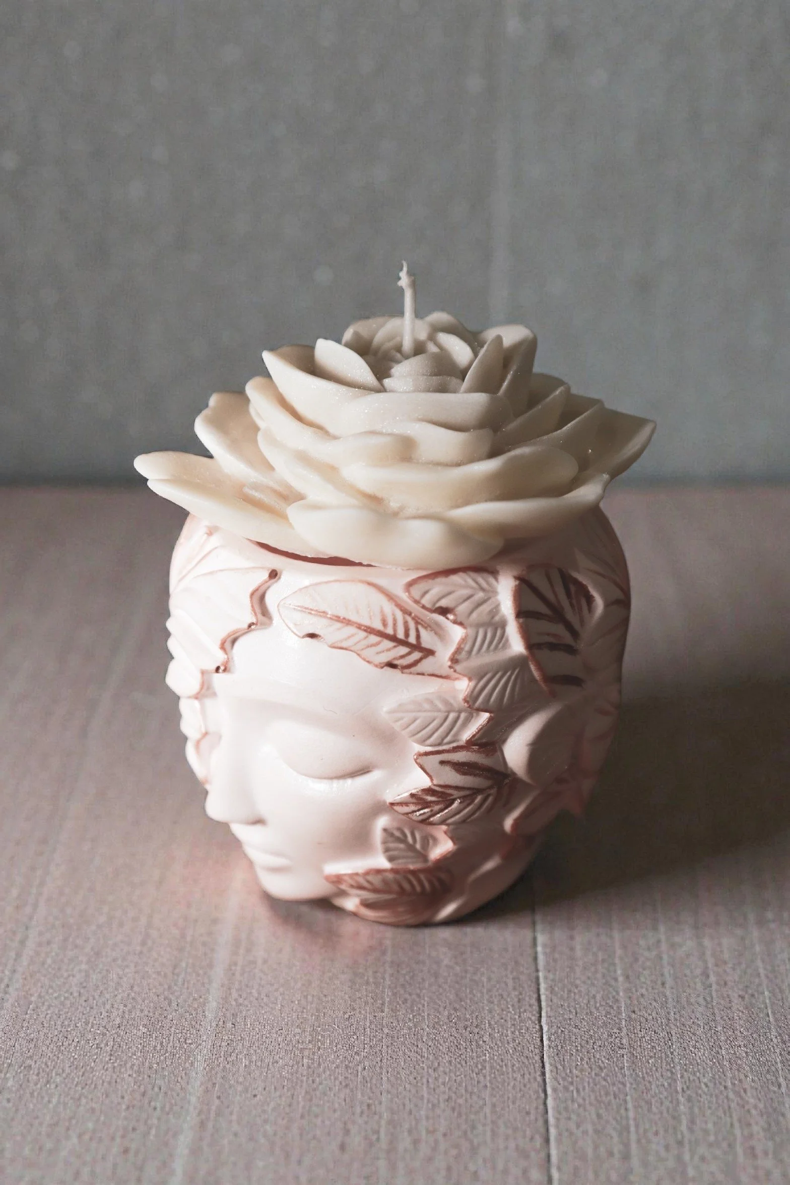 A decorative ceramic vase with a textured leaf pattern, holding a flower-shaped candle with layered petals and a central wick, placed on a tiled surface.