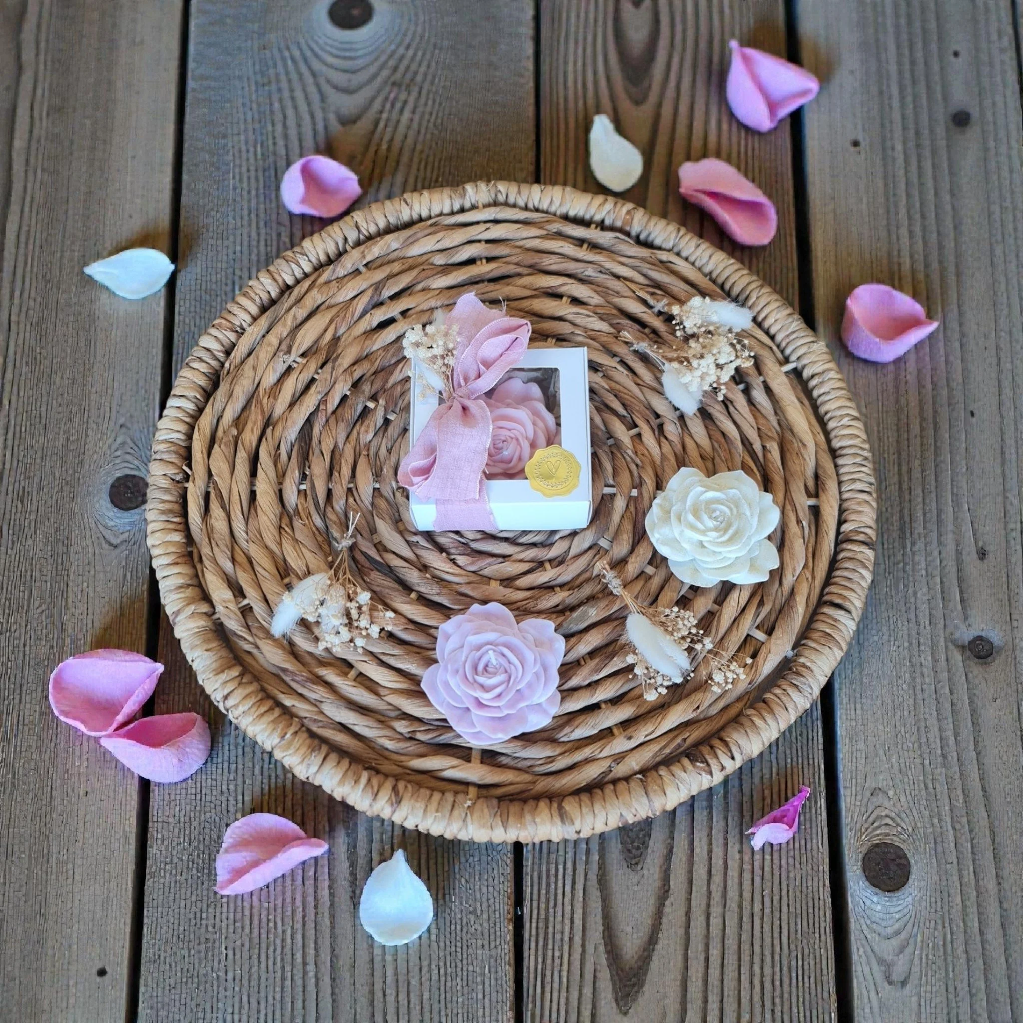 A wicker picnic basket on a wooden surface, surrounded by pink and white flower petals and a printed card with pink fabric roses and a gold seal.