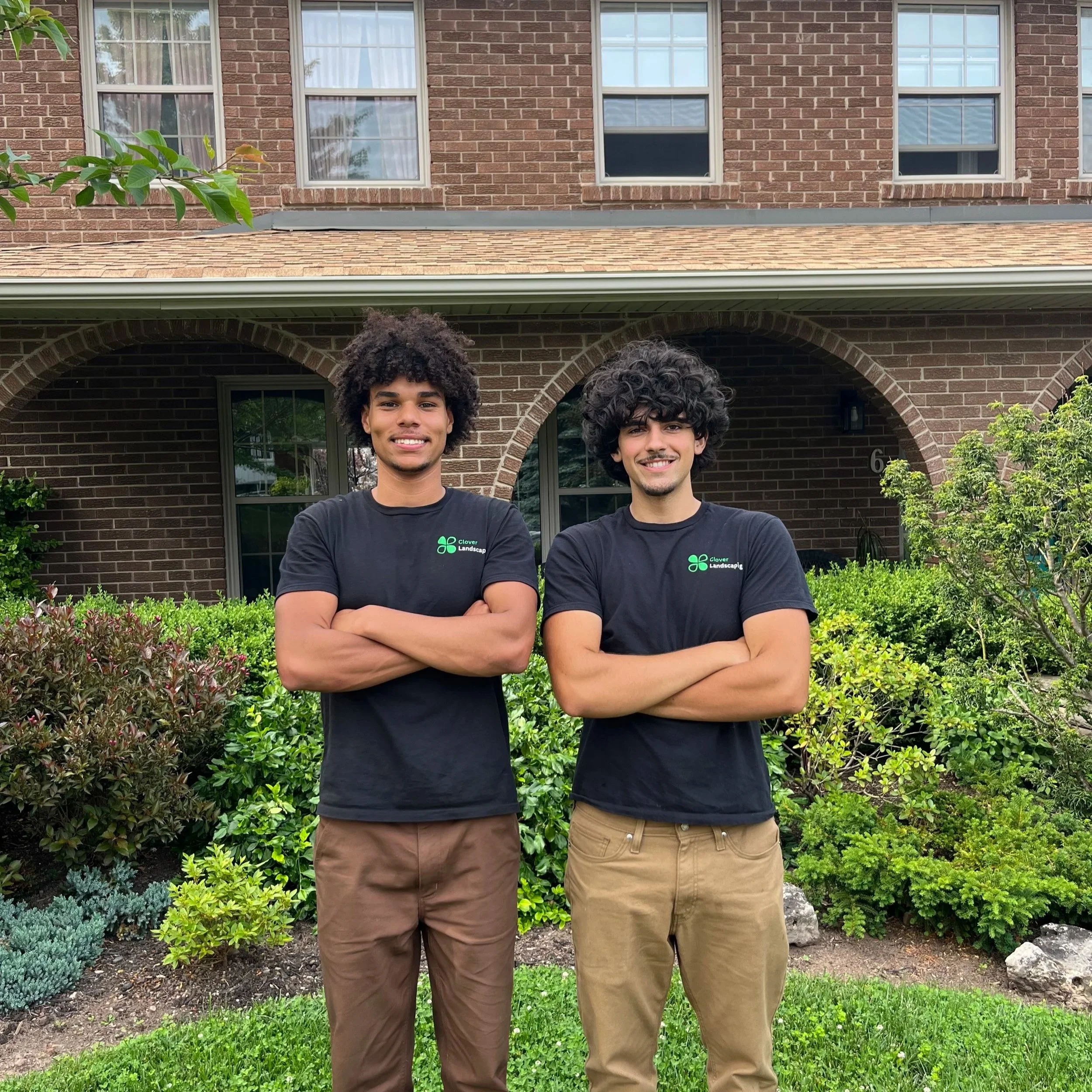 Two young men with crossed arms standing in front of a brick house with arched doorway, greenery, and windows, smiling at the camera.