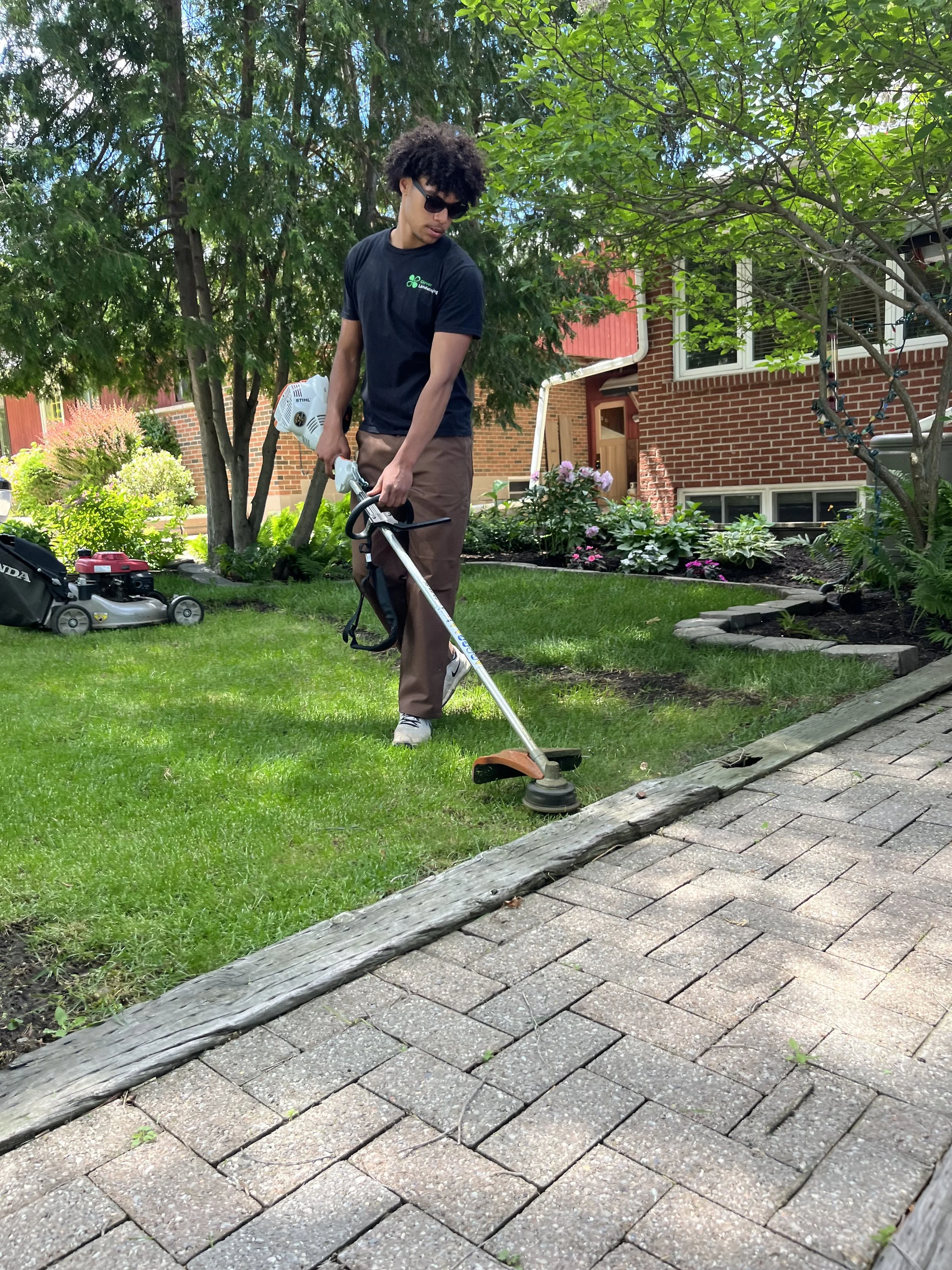 A young man with dark, curly hair, wearing sunglasses, a black t-shirt with a small green logo, and brown pants, is mowing the lawn with a gas-powered string trimmer in a residential yard. There are trees, flowers, and a brick house in the background, with a paved walkway and a wooden log border in the foreground.