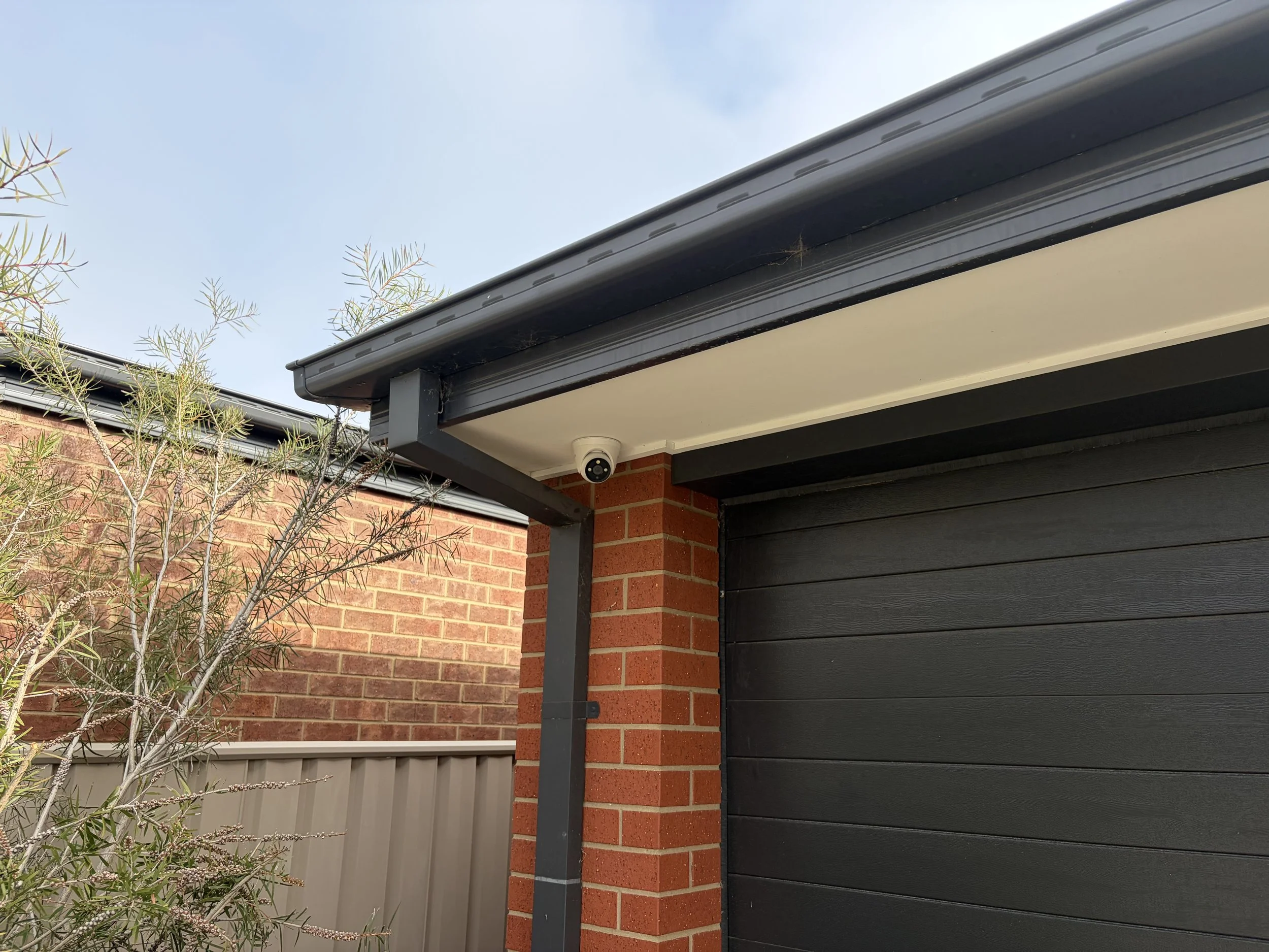 Close-up view of a house corner showing a black metal garage door, a brick wall, a security camera, and tree branches with sparse foliage against a blue sky.