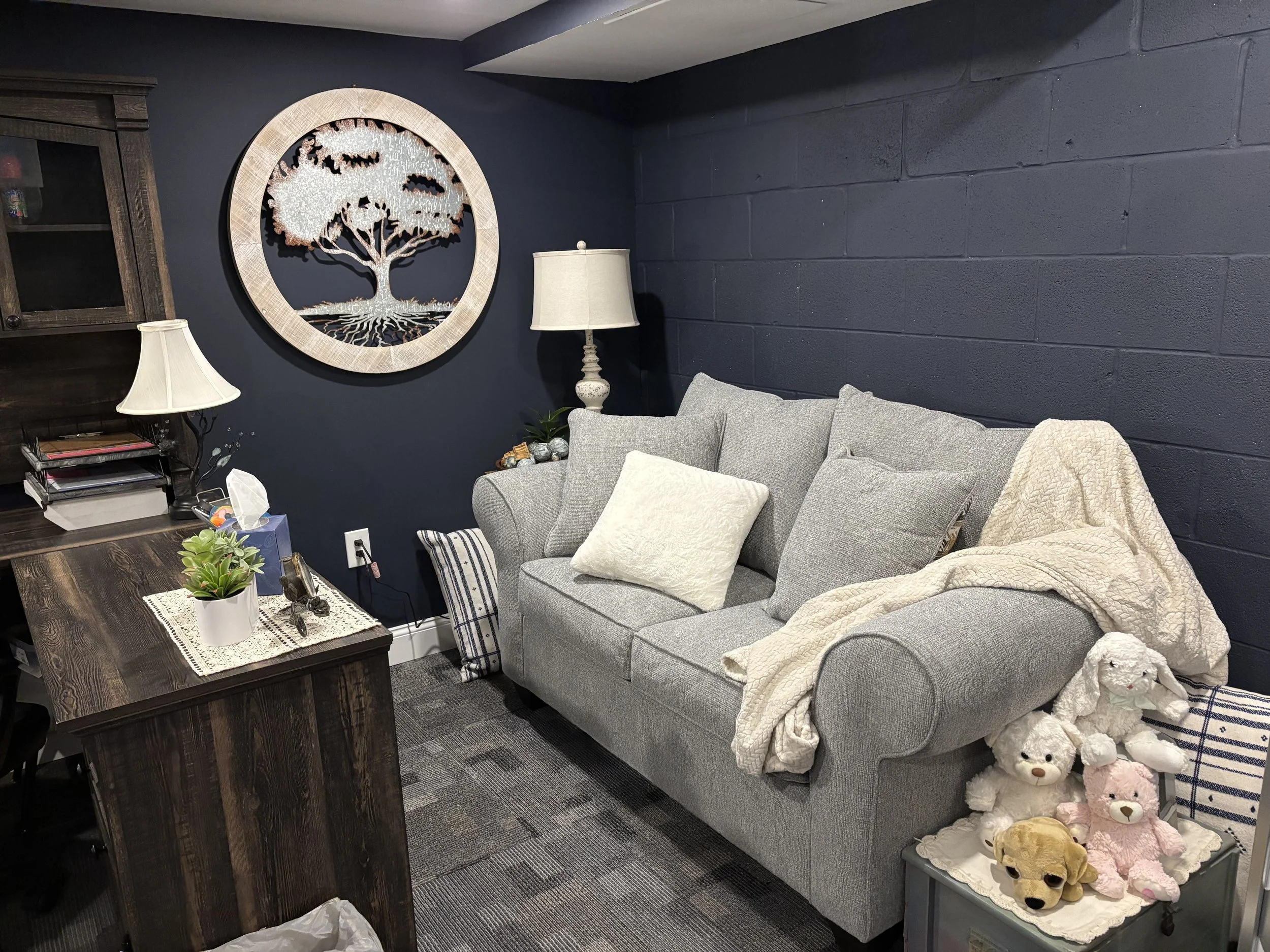 A cozy living room corner with a gray upholstered sofa decorated with cushions, a wooden coffee table with plants and a doily, a standing lamp, a large wall art of a tree with roots, a dark wall, and a shelf with various items including teddy bears and soft toys.