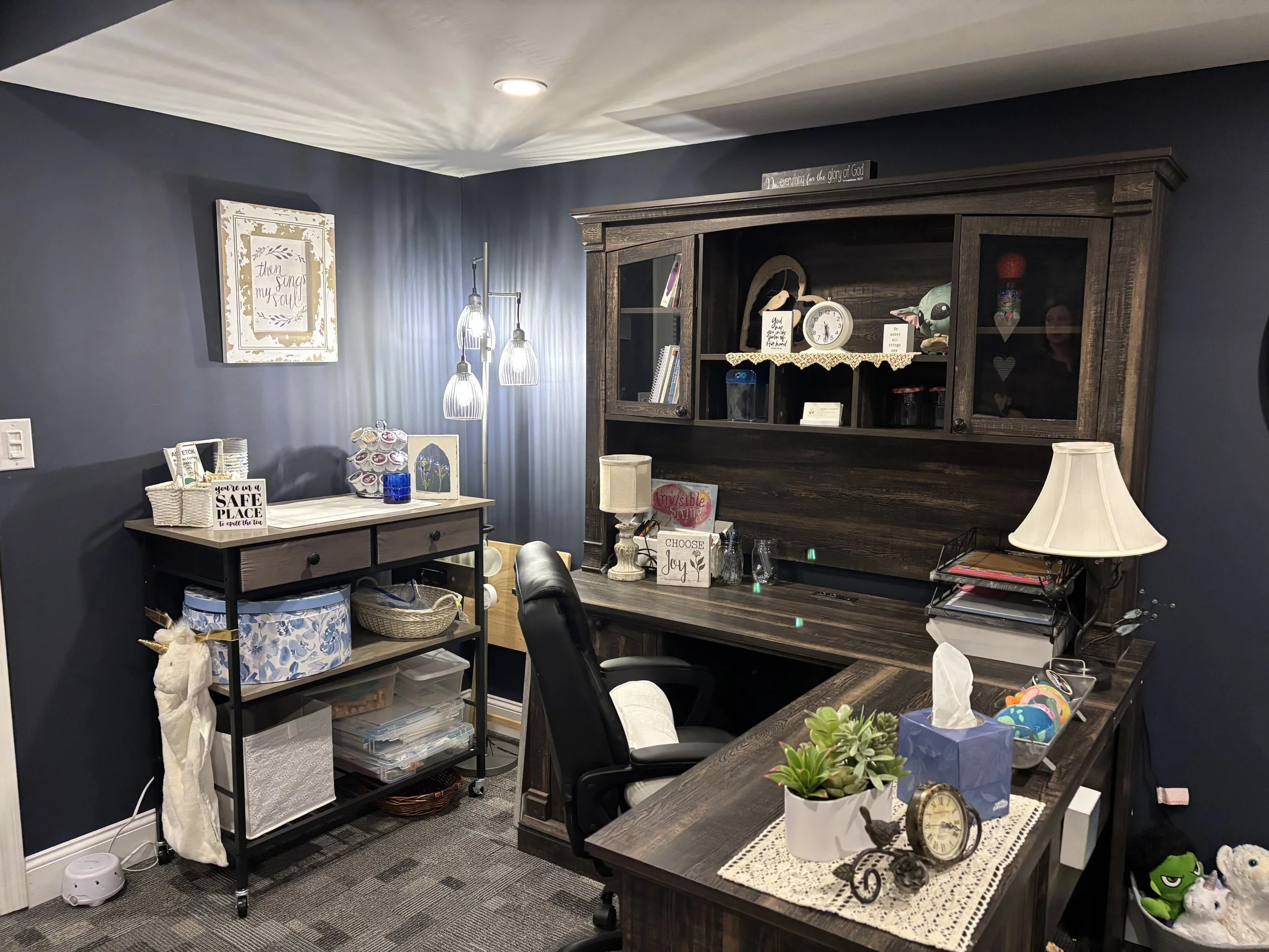 A cozy home office corner with a dark wooden desk and a beige lampshade, a dark wooden hutch with decorative items and books, a small black rolling cart filled with various supplies, and a wall art piece. The room features dark blue walls and gray patterned carpeting.