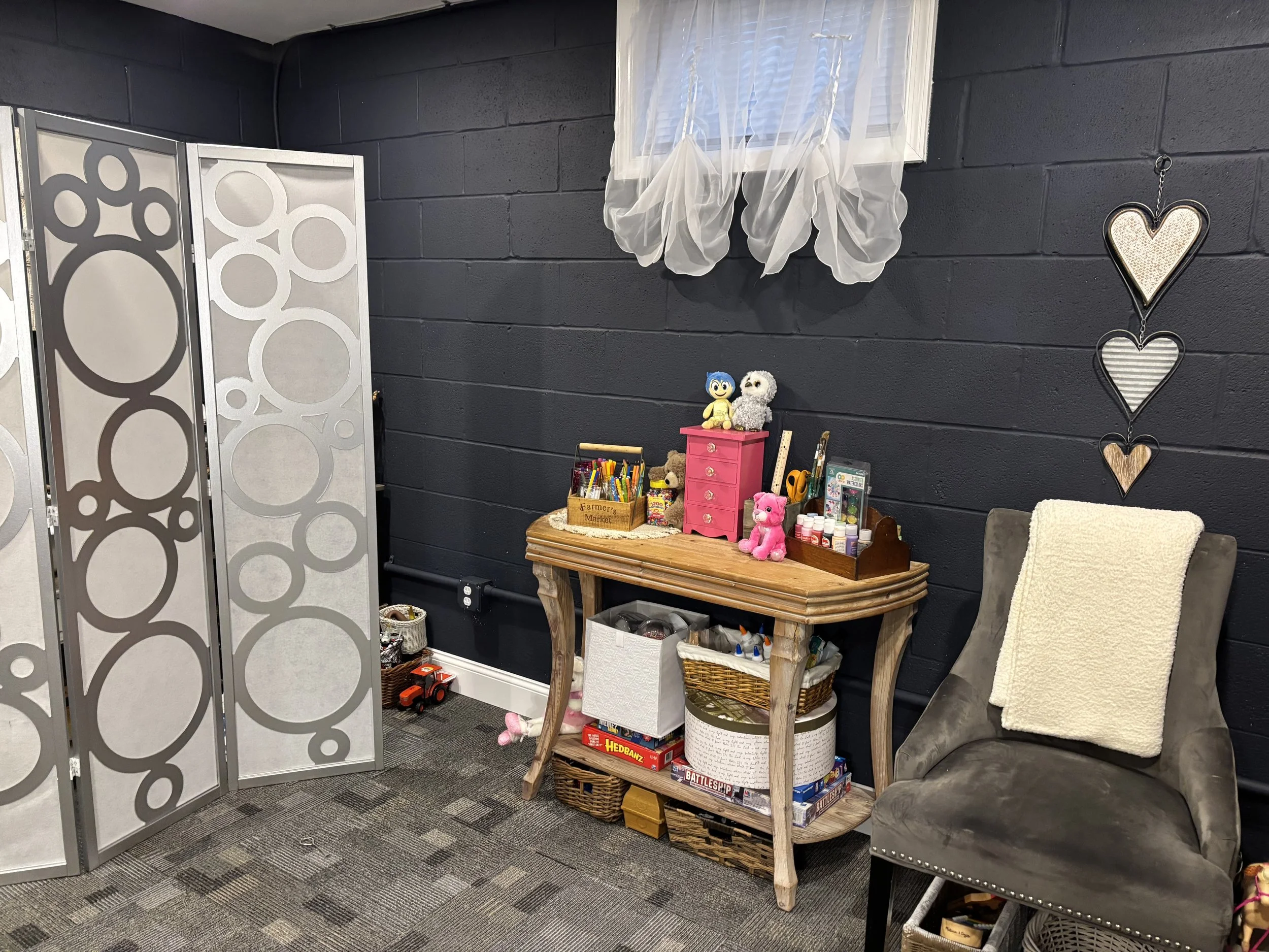 This image shows an indoor space with dark painted brick walls, a window with sheer curtains, and decorative hearts hanging on the wall. There is a small wooden table holding toys, art supplies, and board games. To the right, there's a gray armchair with a white pillow and a beige blanket. A decorative partition with circular cutouts partially separates the space. There are various storage baskets and a small red toy truck on the carpeted floor.