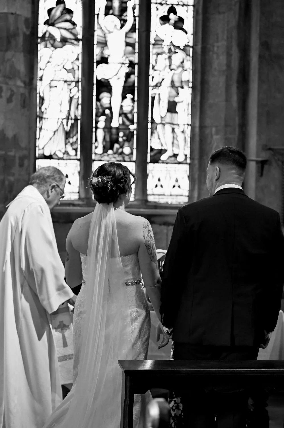 Bride and groom standing at the altar during church wedding ceremony, officiant reading, stunning stained glass windows, black and white documentary wedding photography Nottinghamshire