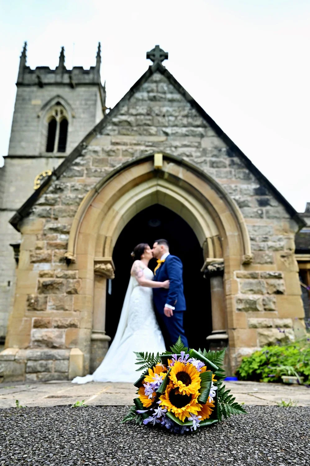 Bride and groom kissing in church doorway with sunflower bouquet in foreground, creative wedding portrait at historic stone church, Doncaster wedding photographer