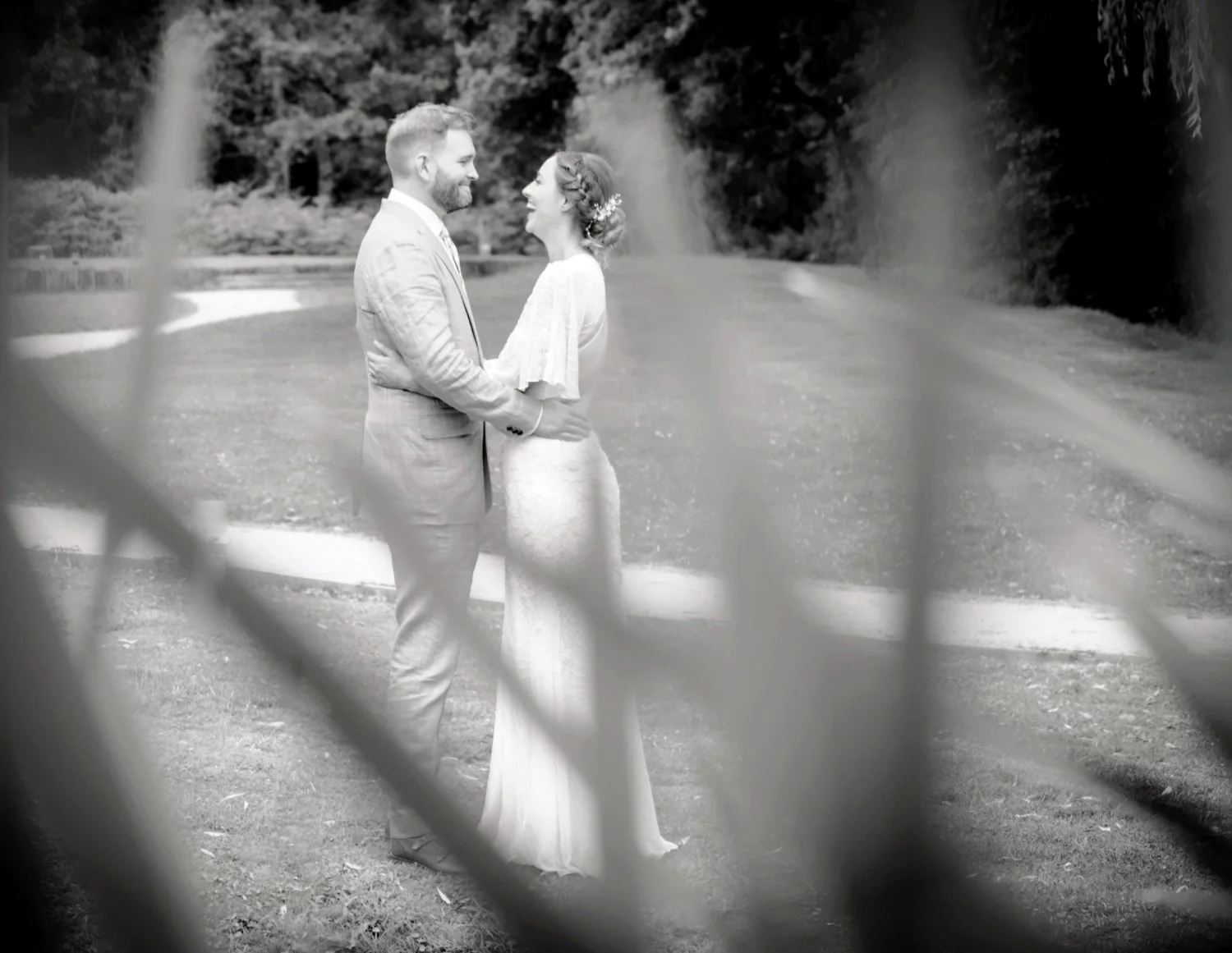 Black and white bride and groom portrait shot through pond reeds across water, couple laughing in garden grounds, creative documentary wedding photography Nottinghamshire