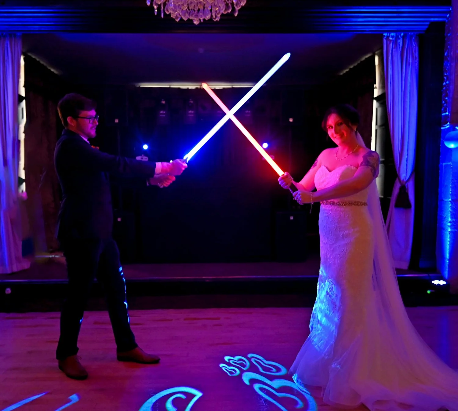 A bride and groom in formal attire holding illuminated lightsabers and posing for a photo during a reception or celebration.