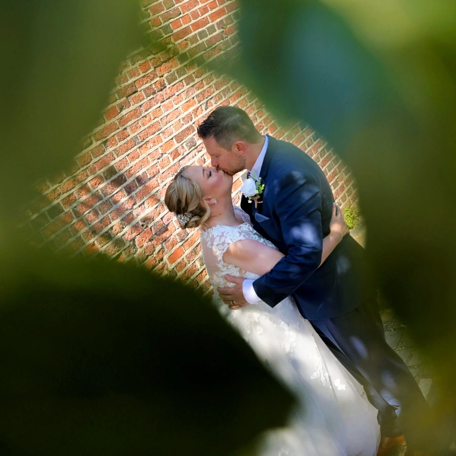 Bride and groom sharing a romantic kiss framed through garden foliage against a brick wall — creative natural framing wedding photography by Tasha Boorman, Nottinghamshire wedding photographer