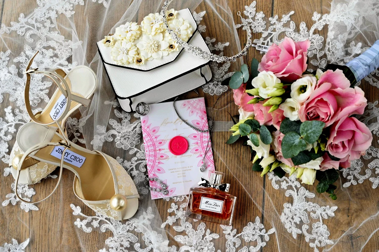 Wedding detail flatlay with Jimmy Choo heels, Miss Dior perfume, pink wax seal invitation, floral clutch bag, diamond tiara and pink rose bouquet on lace veil, luxury wedding detail photography Chewton Glen Hampshire