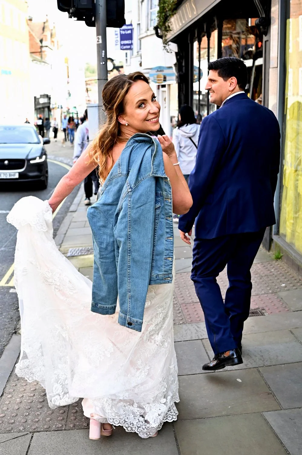 Bride in wedding dress and denim jacket looking back with a cheeky smile while walking through Leicester city centre streets — urban wedding photography by Tasha Boorman, Leicestershire wedding photographer