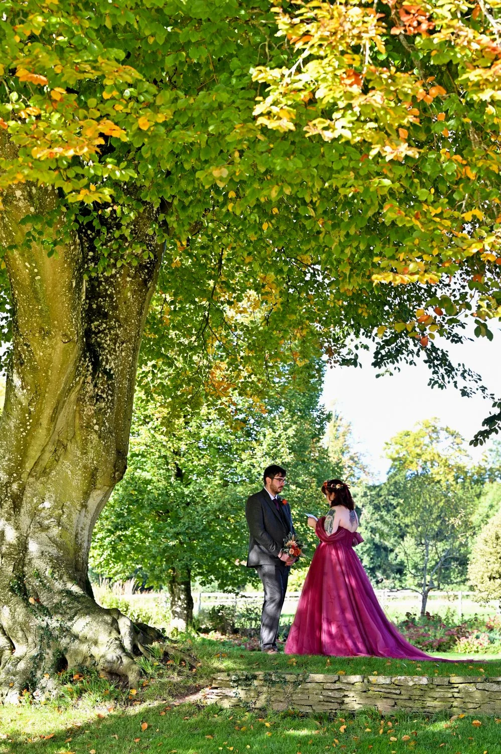 Bride in a stunning pink ballgown reading personal vows to her groom beneath a magnificent autumn tree — alternative wedding photography by Tasha Boorman, Nottinghamshire wedding photographer