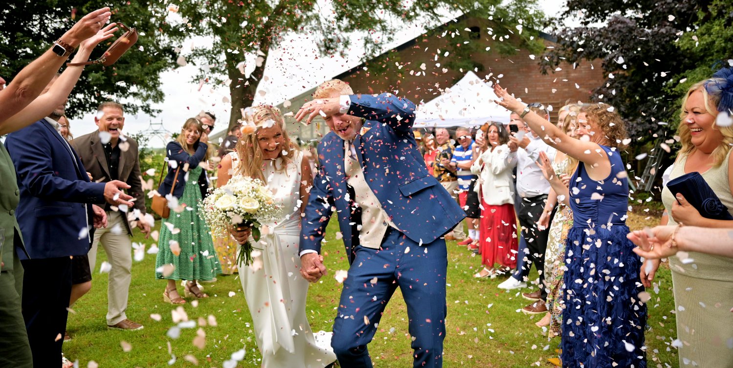 Bride and groom walking through confetti shower after wedding ceremony, groom shielding face laughing, guests cheering, candid documentary wedding photography Leicestershire farm wedding venue