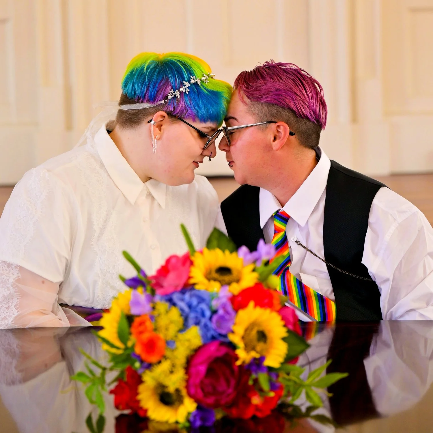 Two people with rainbow-colored hair, glasses, and wedding attire touching foreheads at a wedding ceremony, with a vibrant flower bouquet in the foreground.