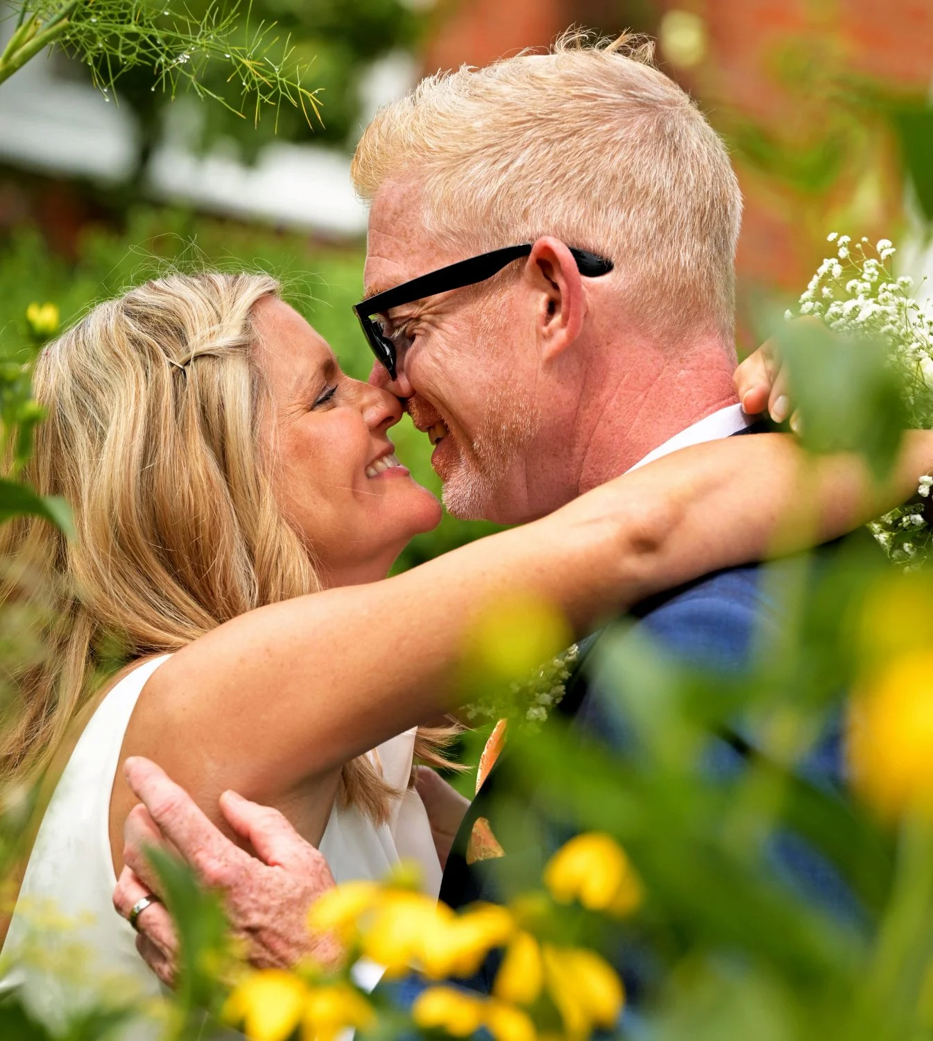 Bride and groom laughing and almost kissing, framed by yellow wildflowers and greenery, candid outdoor wedding portrait at farm wedding venue, Leicestershire wedding photographer